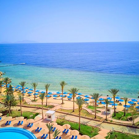 Pickalbatros Royal Grand Sharm - Adults Friendly 16 Years Plus Exteriér fotografie The photo displays a beautiful coastal scene featuring a clear blue sea that transitions into green tones near the shore. In the foreground, there are sandy beaches lined with palm trees and a variety of beach umbrellas. There is also a swimming pool