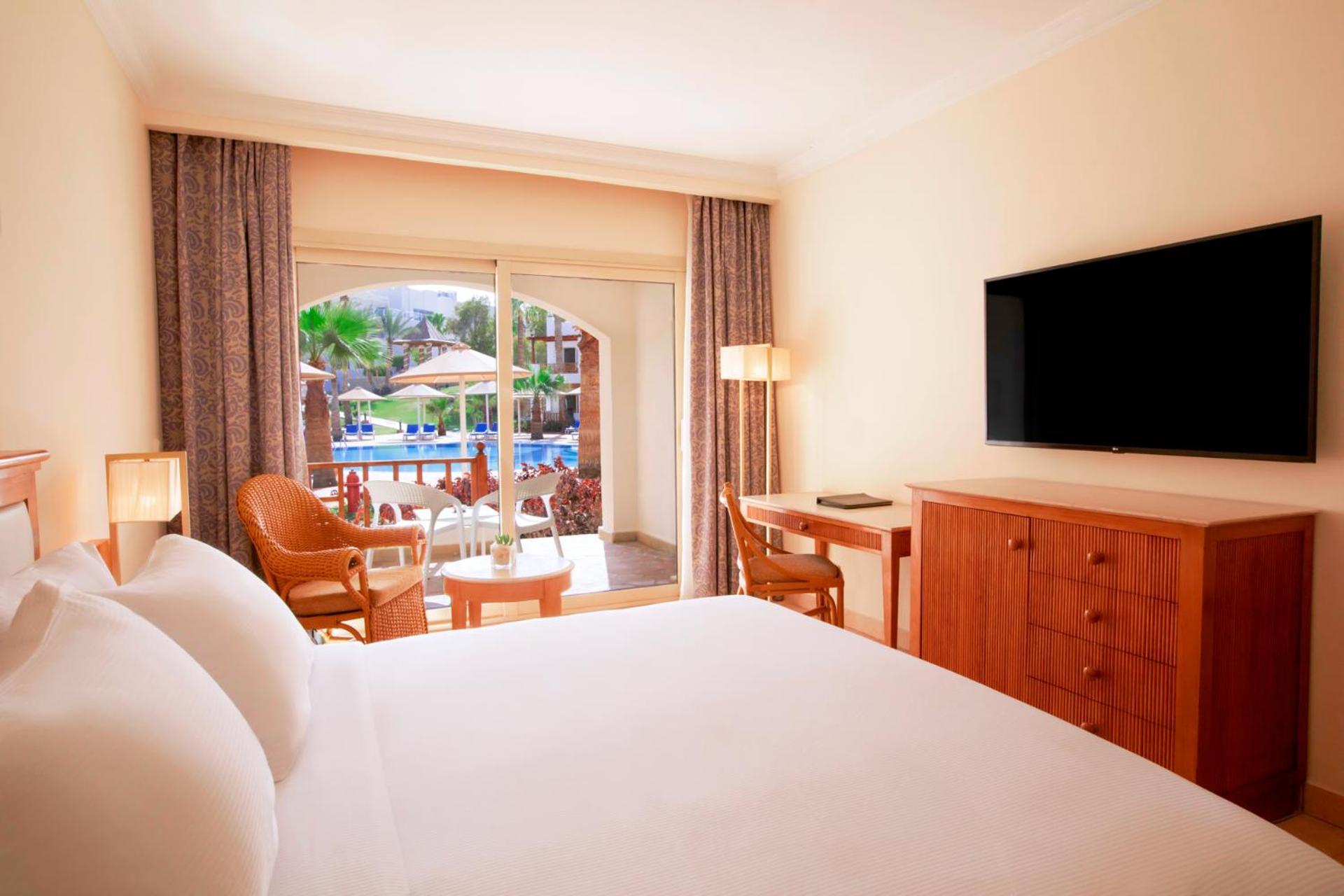Pickalbatros Royal Grand Sharm - Adults Friendly 16 Years Plus Exteriér fotografie The photo shows a hotel room featuring a large bed with white bedding. There are wooden furniture pieces, including a dresser and a desk. A flat-screen television is mounted on the wall opposite the bed. The room has large windows with sheer curtains