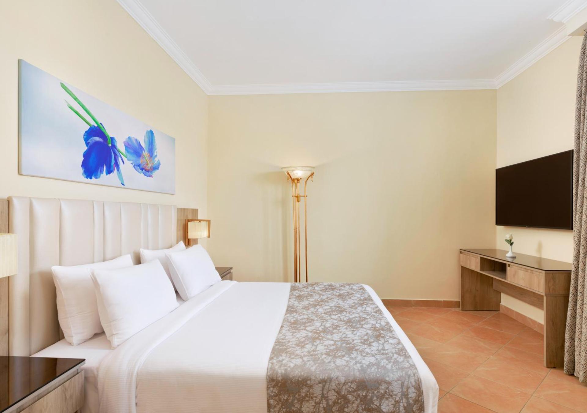 Pickalbatros Royal Grand Sharm - Adults Friendly 16 Years Plus Exteriér fotografie The photo shows a modern hotel room featuring a large bed with white linens and a decorative gray throw. On one side of the room, there is a piece of artwork depicting blue flowers. A floor lamp provides additional lighting, and across from the bed i
