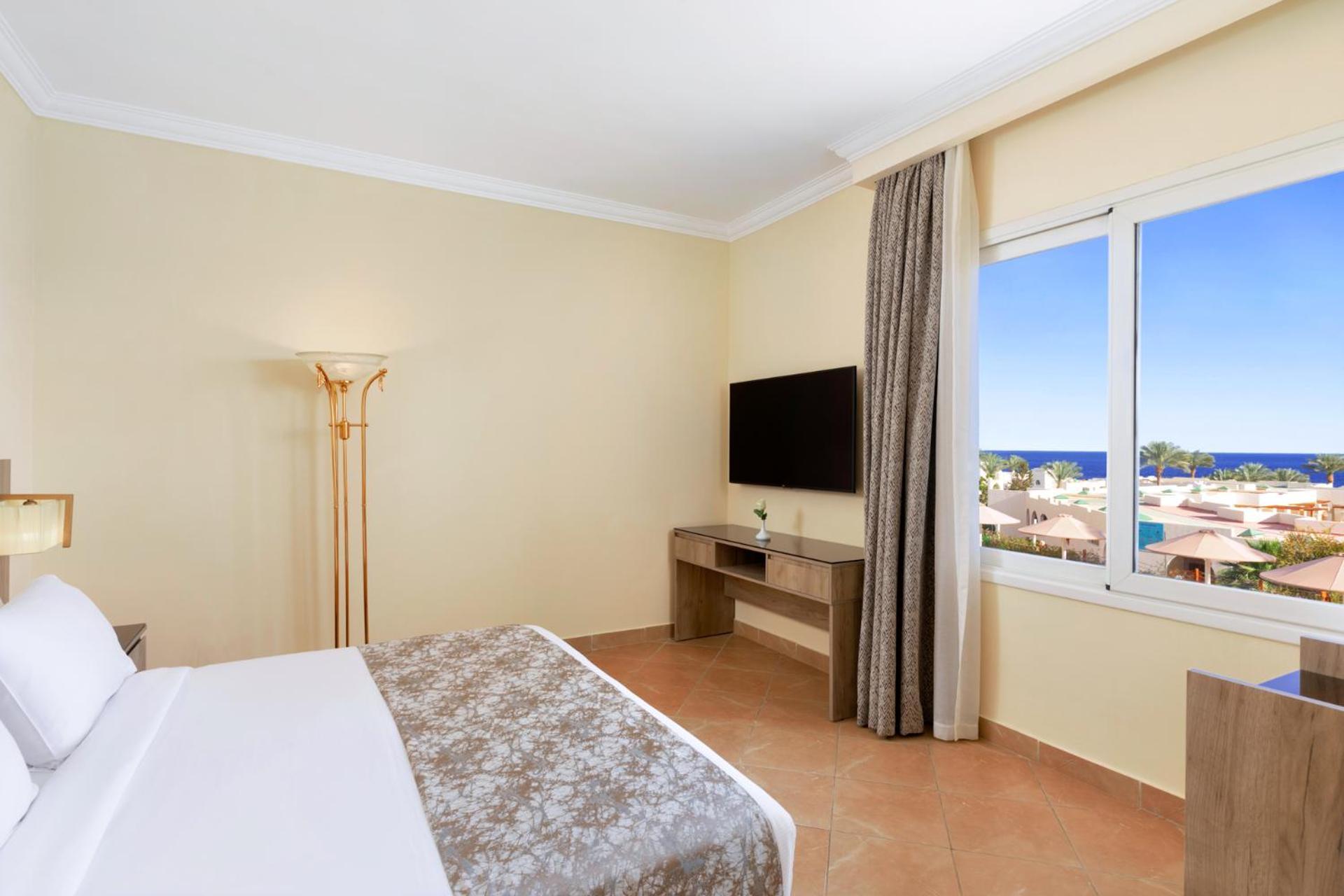 Pickalbatros Royal Grand Sharm - Adults Friendly 16 Years Plus Exteriér fotografie The photo shows a hotel room with a modern and minimalistic design. There is a comfortable bed with white linens and a decorative gray throw at the foot. A large window offers a view of the outdoors, possibly with a glimpse of the sea or beach. A fla