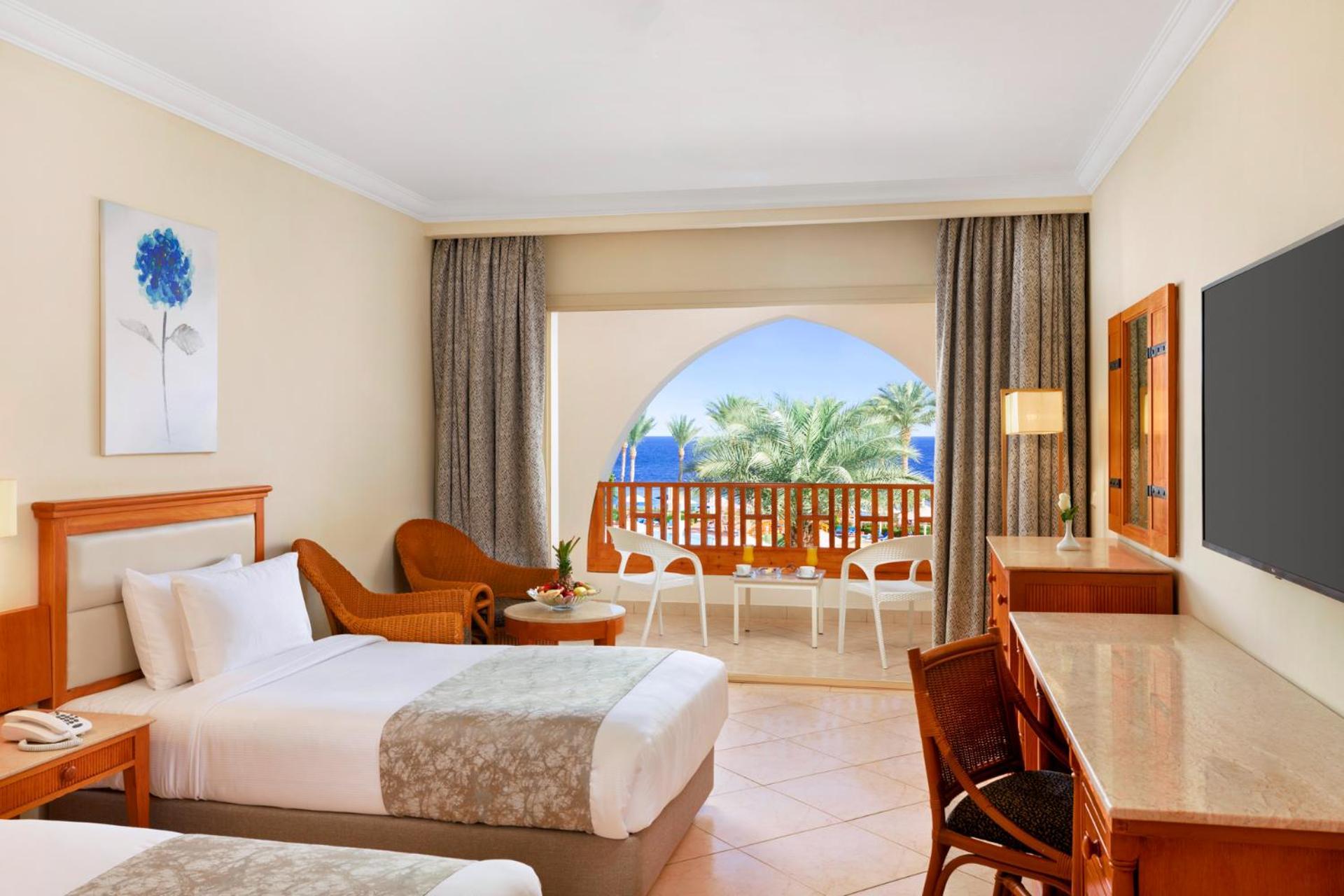 Pickalbatros Royal Grand Sharm - Adults Friendly 16 Years Plus Exteriér fotografie The photo shows a hotel room featuring two beds with light bedding. There is a wooden desk and chair, and a comfortable chair is placed nearby. The room has large windows that open to a balcony with a view of palm trees and a blue sky. The decor is r