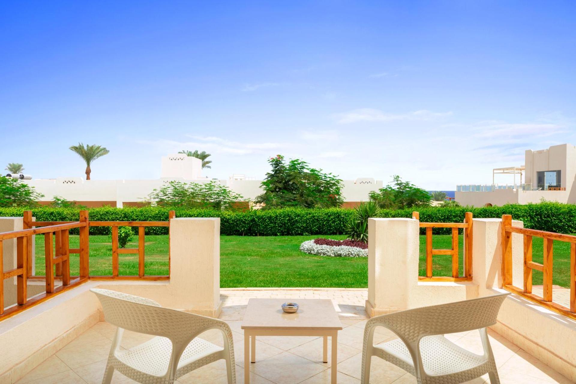 Pickalbatros Royal Grand Sharm - Adults Friendly 16 Years Plus Exteriér fotografie The photo depicts a serene outdoor balcony or patio area. It features two white chairs and a small table, set against a backdrop of lush green grass and garden plants. In the distance, there are some palm trees and clear blue skies, suggesting a warm