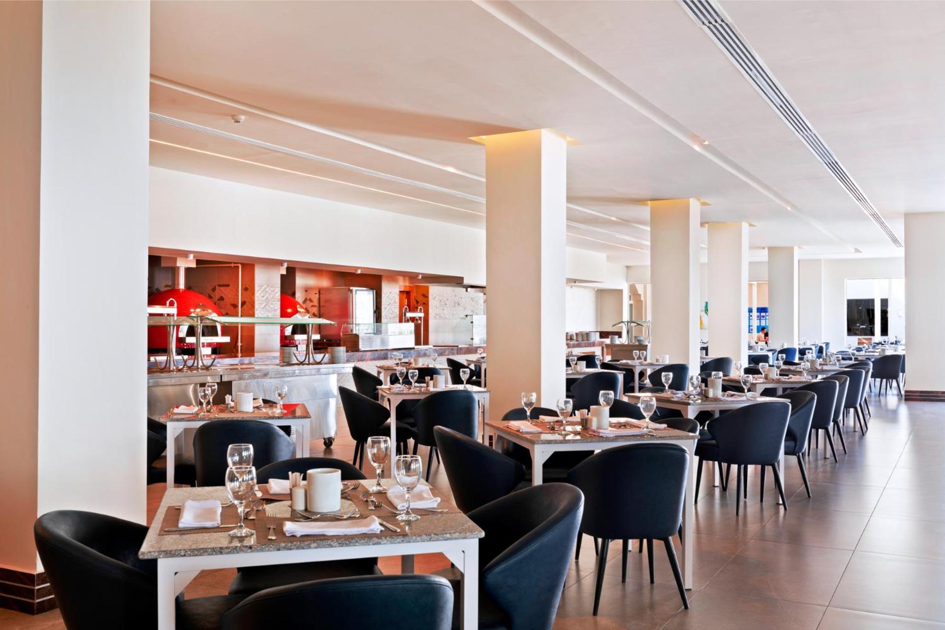 Pickalbatros Royal Grand Sharm - Adults Friendly 16 Years Plus Exteriér fotografie The photo shows a modern restaurant interior. It features a spacious layout with several dining tables arranged in a row, each set with dishes, glasses, and cutlery. The tables have light-colored surfaces and are accompanied by black chairs. In the b