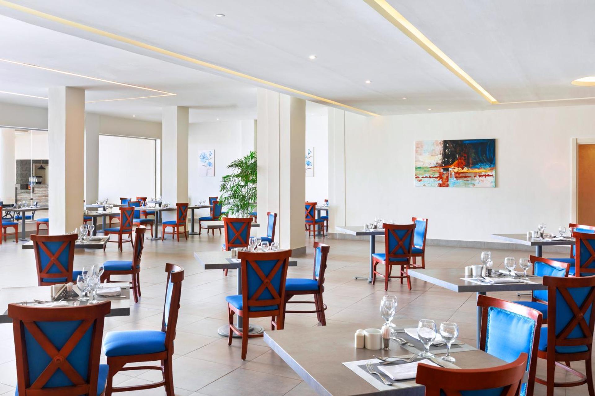 Pickalbatros Royal Grand Sharm - Adults Friendly 16 Years Plus Exteriér fotografie The photo shows an interior of a restaurant featuring a modern and spacious design. The floors are tiled, and the walls are painted in neutral tones. The seating consists of wooden chairs with blue cushions arranged around tables, which are set with 