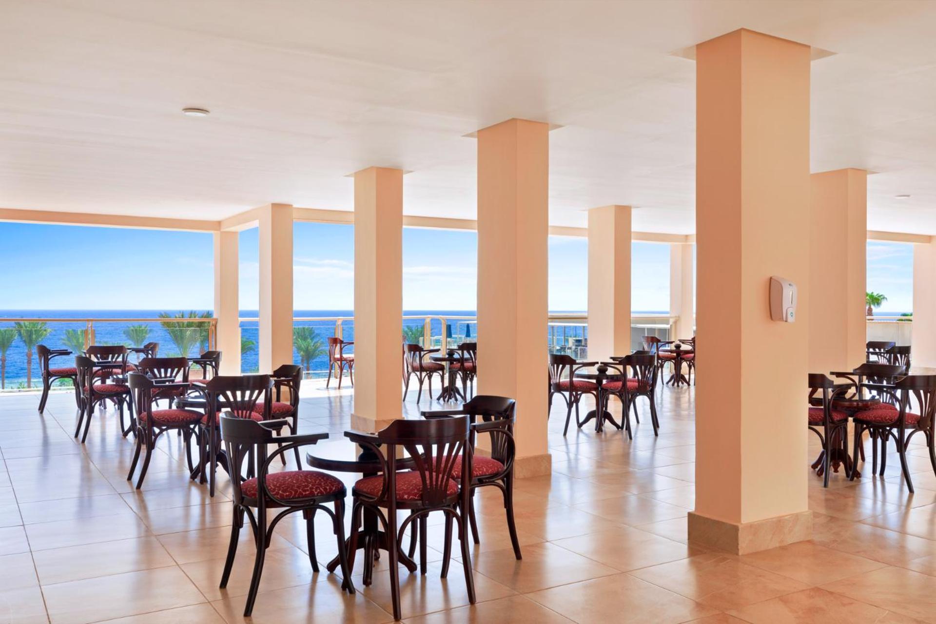 Pickalbatros Royal Grand Sharm - Adults Friendly 16 Years Plus Exteriér fotografie The image shows a dining area or café with several round wooden tables and chairs, featuring red upholstery. The space is well-lit, with large windows that allow a view of the ocean in the background, contributing to a bright and open atmosphere. The