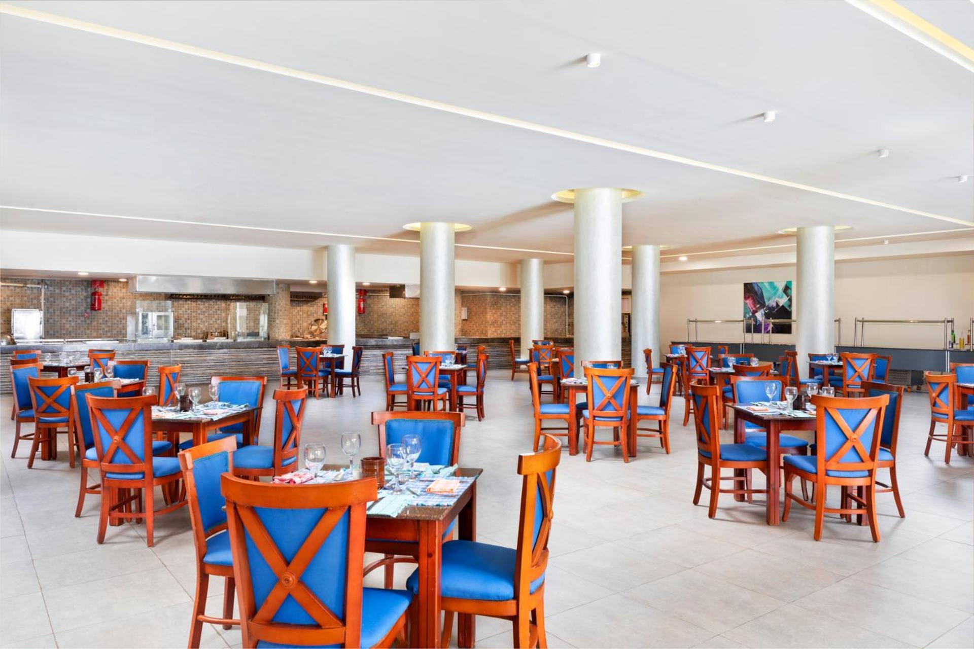 Pickalbatros Royal Grand Sharm - Adults Friendly 16 Years Plus Exteriér fotografie The photo depicts a spacious dining area in a restaurant. The room features several wooden tables, each with blue chairs. The chairs have a unique design with prominent X-shaped backs. The flooring is light-colored tile, and the walls are painted in 