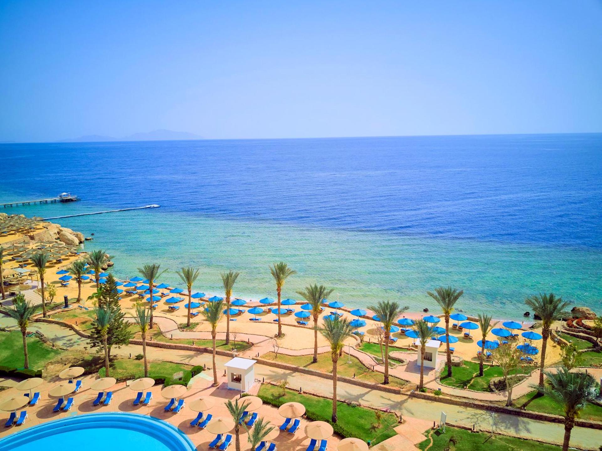Pickalbatros Royal Grand Sharm - Adults Friendly 16 Years Plus Exteriér fotografie The photo displays a beautiful coastal scene featuring a clear blue sea that transitions into green tones near the shore. In the foreground, there are sandy beaches lined with palm trees and a variety of beach umbrellas. There is also a swimming pool