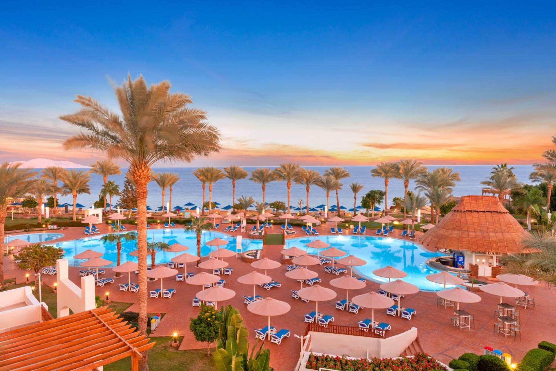 Pickalbatros Royal Grand Sharm - Adults Friendly 16 Years Plus Exteriér fotografie The photo depicts a scenic view of a resort area featuring a large swimming pool surrounded by numerous lounge chairs and umbrellas. In the background, there are tall palm trees and a beautiful sunset over the horizon, creating a vibrant sky. The atm