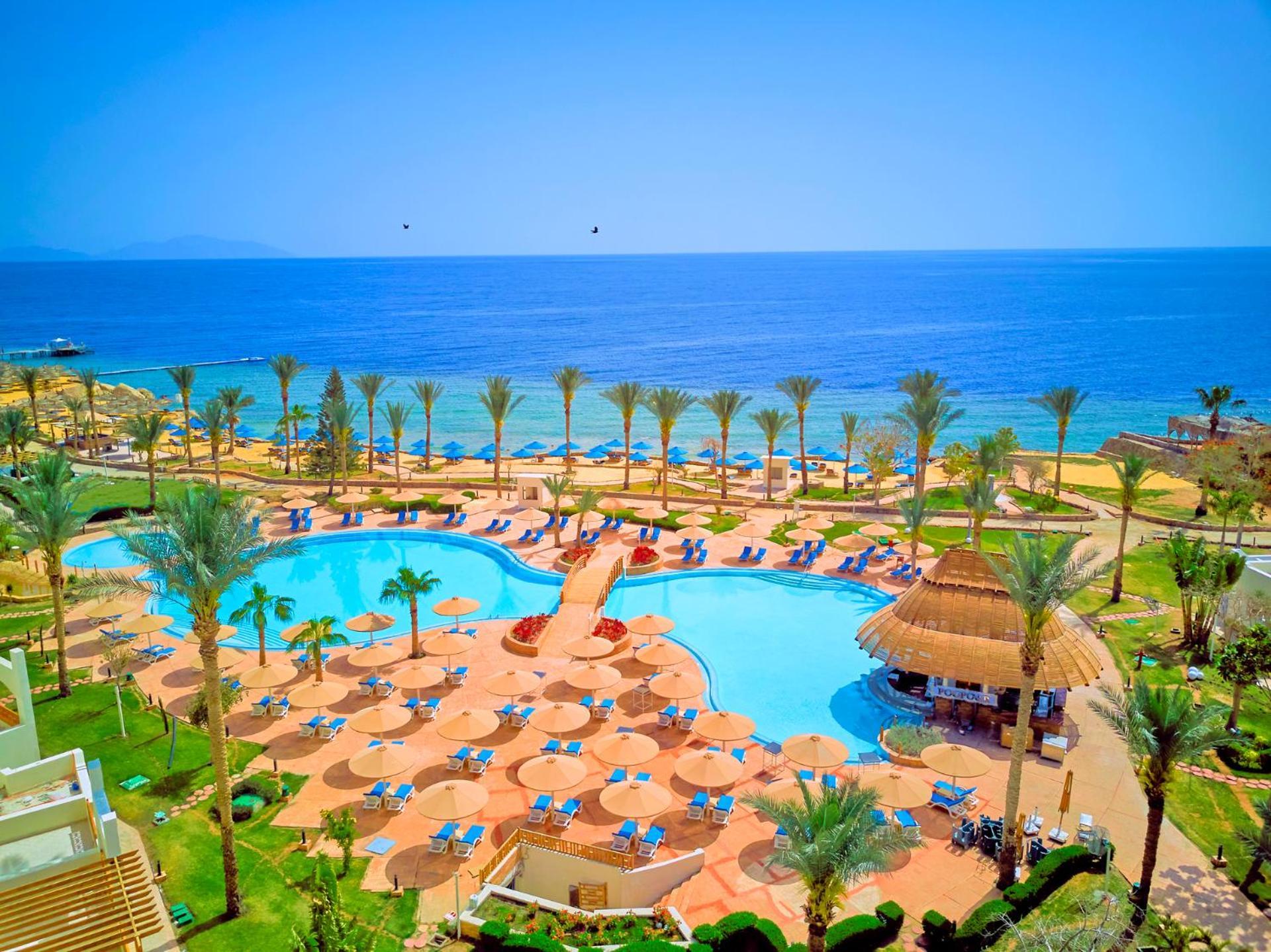 Pickalbatros Royal Grand Sharm - Adults Friendly 16 Years Plus Exteriér fotografie The photo depicts a picturesque resort scene by the sea. In the foreground, there is a large, inviting swimming pool surrounded by lounge chairs and umbrellas. Palm trees are strategically placed around the pool area, adding to the tropical atmospher