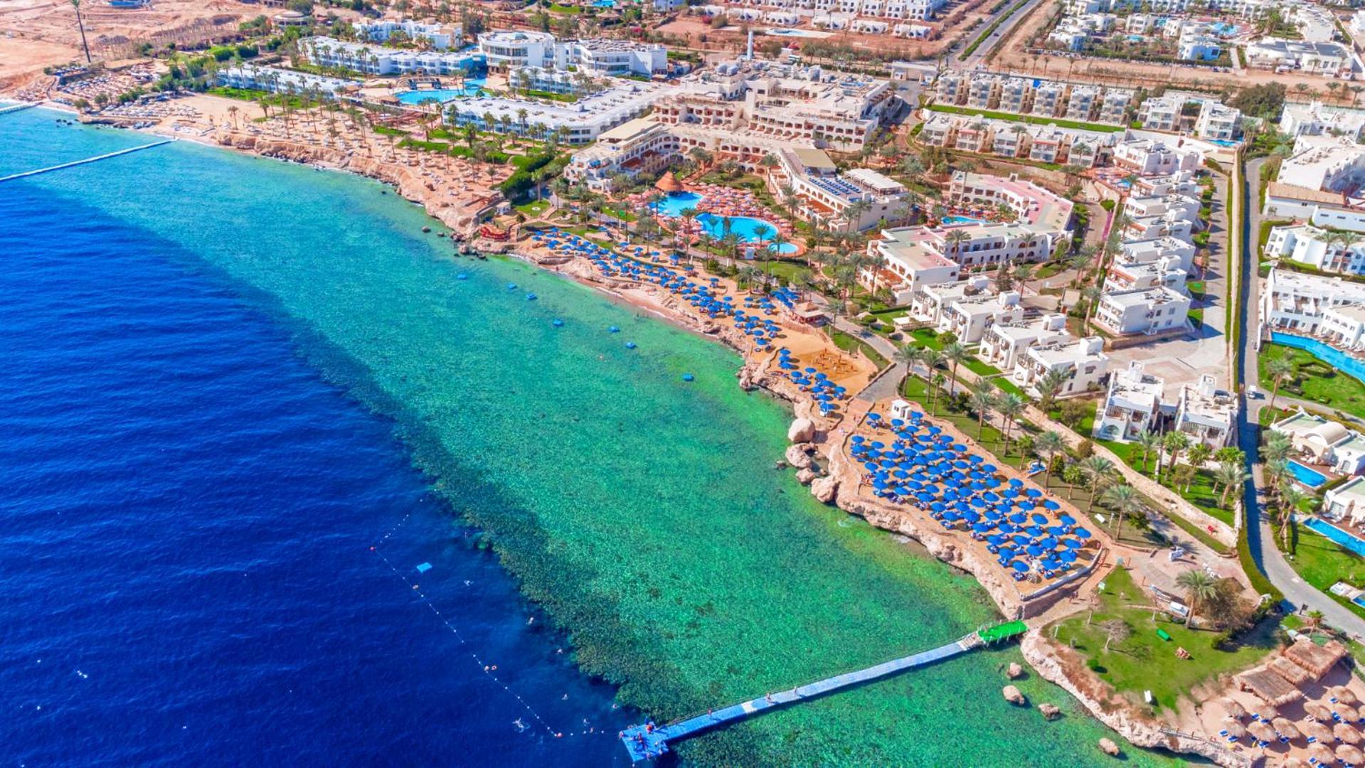Pickalbatros Royal Grand Sharm - Adults Friendly 16 Years Plus Exteriér fotografie The photo showcases a beautiful aerial view of a coastal resort. In the foreground, there's a sandy beach with beach umbrellas and lounge chairs arranged neatly, indicating a tourist area. The clear, turquoise water transitions from the sandy shore t