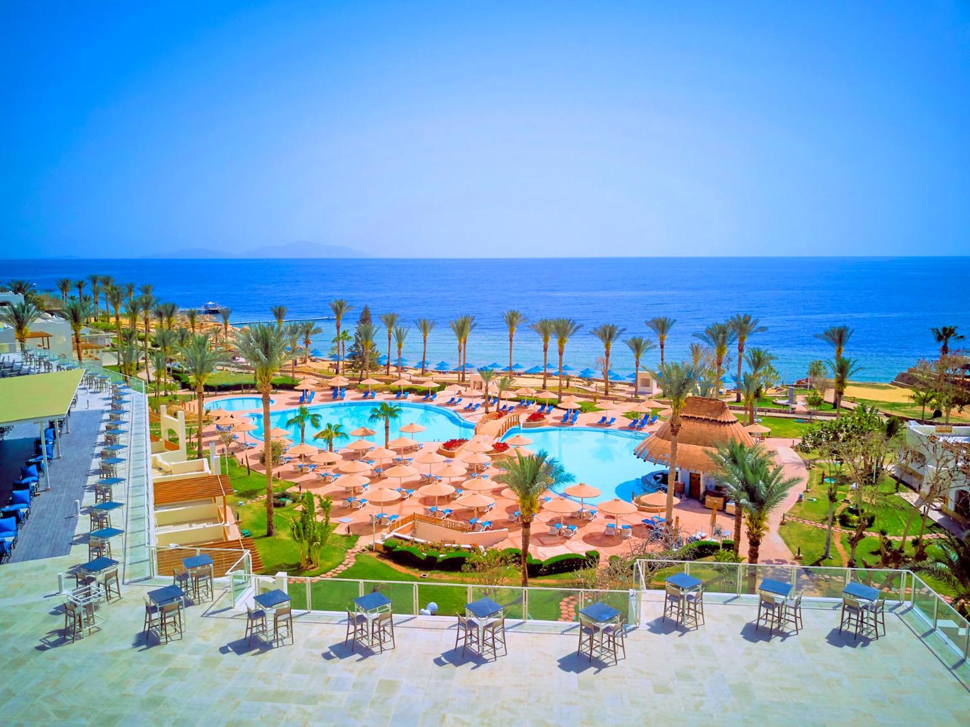Pickalbatros Royal Grand Sharm - Adults Friendly 16 Years Plus Exteriér fotografie The photo shows a beautiful resort scene with a spacious swimming pool at the center, surrounded by sun loungers and palm trees. There are areas for relaxation with tables and chairs positioned around the pool. In the background, the vibrant blue oce