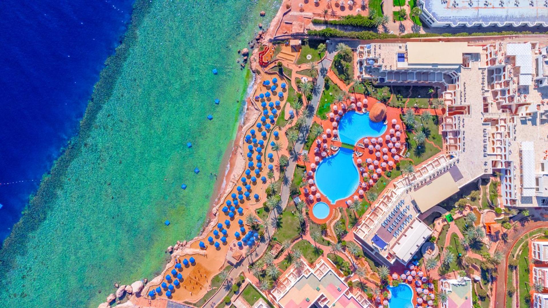 Pickalbatros Royal Grand Sharm - Adults Friendly 16 Years Plus Exteriér fotografie The photo showcases an aerial view of a resort area by the sea. It features a turquoise body of water alongside a sandy beach. There are several umbrellas set up on the beach and lounging areas. A large pool is visible, characterized by its unique sh