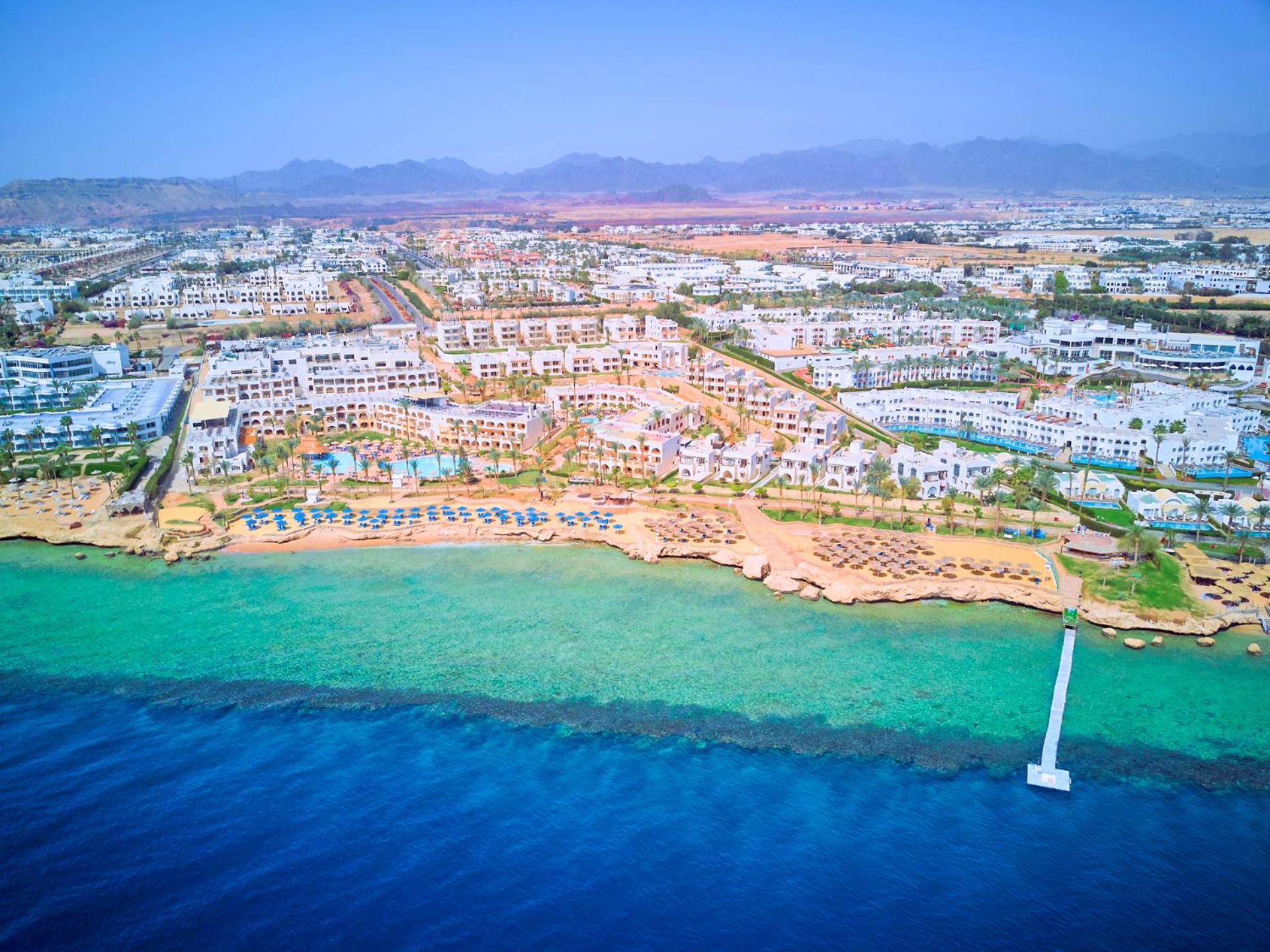 Pickalbatros Royal Grand Sharm - Adults Friendly 16 Years Plus Exteriér fotografie The photo shows an aerial view of a coastal resort area. In the foreground, there is a clear blue ocean with a gradient of colors from deep blue to lighter shades near the shore. The beach features sandy areas lined with sun loungers and umbrellas. F