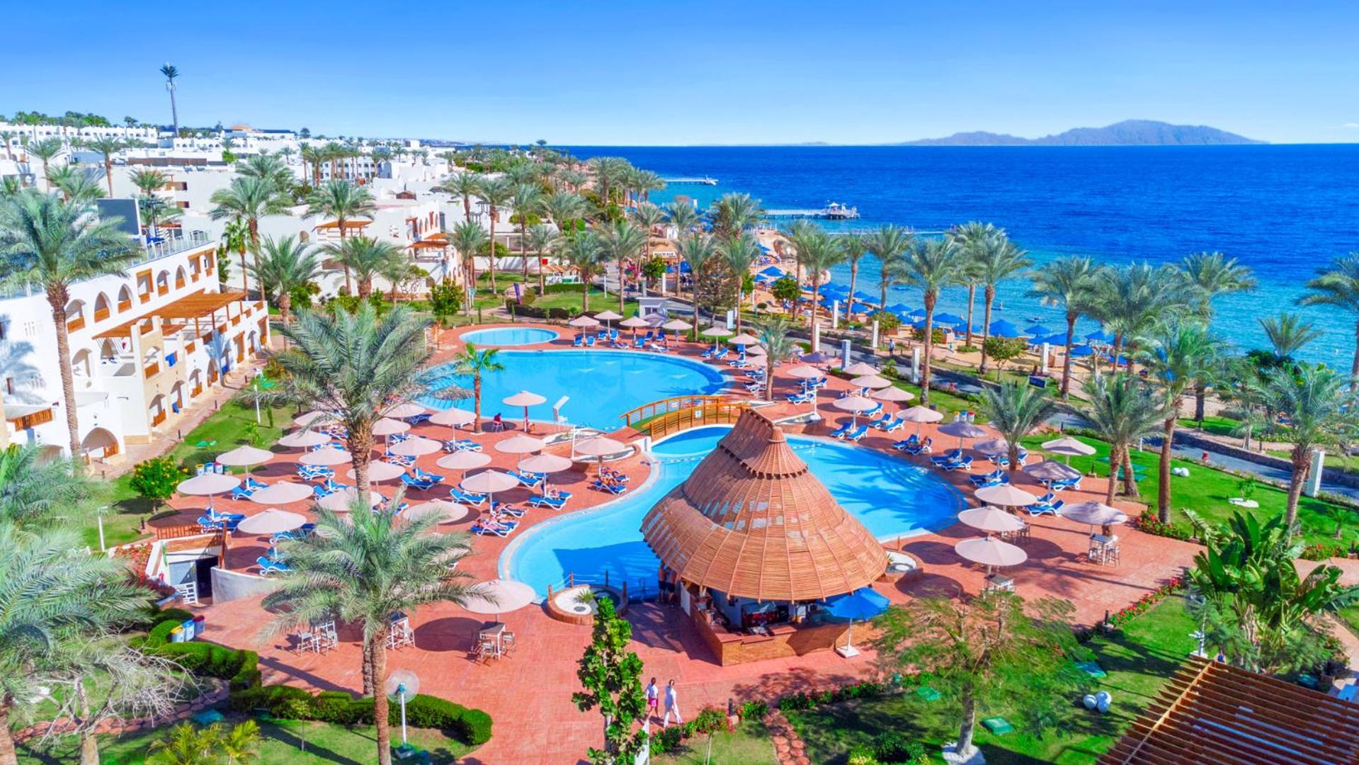 Pickalbatros Royal Grand Sharm - Adults Friendly 16 Years Plus Exteriér fotografie The image depicts a vibrant resort scene overlooking a clear blue sea. In the foreground, there are multiple swimming pools surrounded by sun loungers and umbrellas. A thatched-roof gazebo is situated near the center of the pool area. Palms and lush 