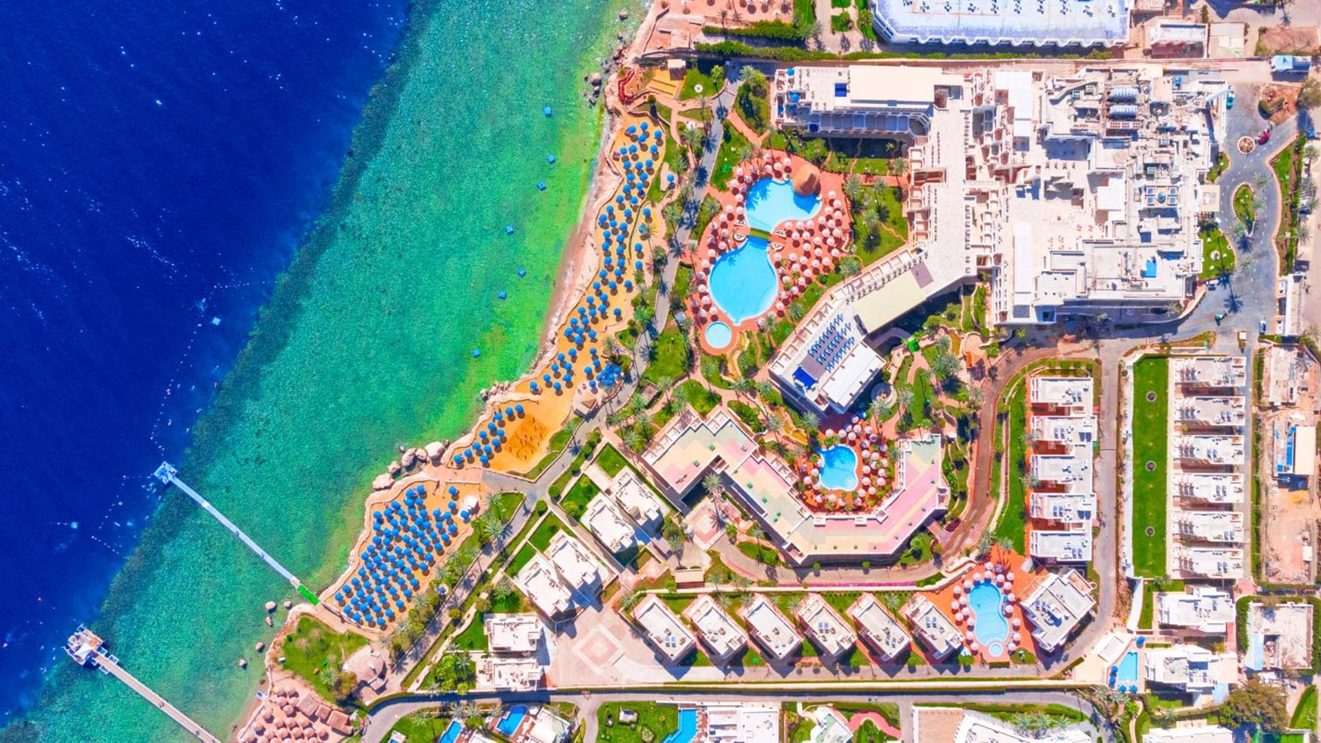 Pickalbatros Royal Grand Sharm - Adults Friendly 16 Years Plus Exteriér fotografie The image shows an aerial view of a coastal resort area. It features various swimming pools outlined by landscaped gardens and pathways. There are also rows of sun loungers and umbrellas near the beach. The water transitions from a deep blue to a vib
