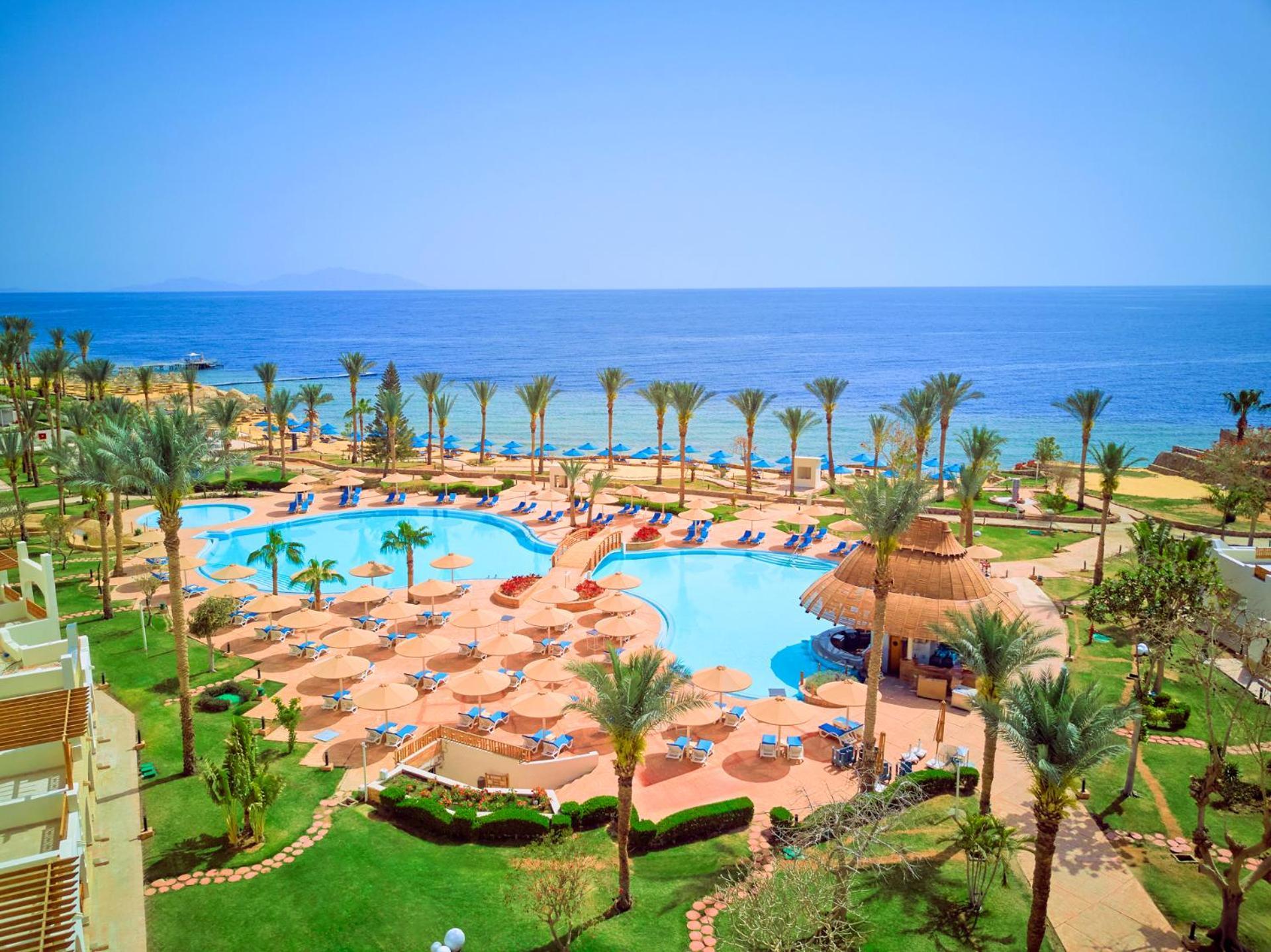 Pickalbatros Royal Grand Sharm - Adults Friendly 16 Years Plus Exteriér fotografie The photo depicts a beautiful resort scene by the ocean. In the foreground, there is a large swimming pool surrounded by palm trees and lounge chairs. The pool area features sunbeds and umbrellas, suggesting a relaxing atmosphere for guests. In the b