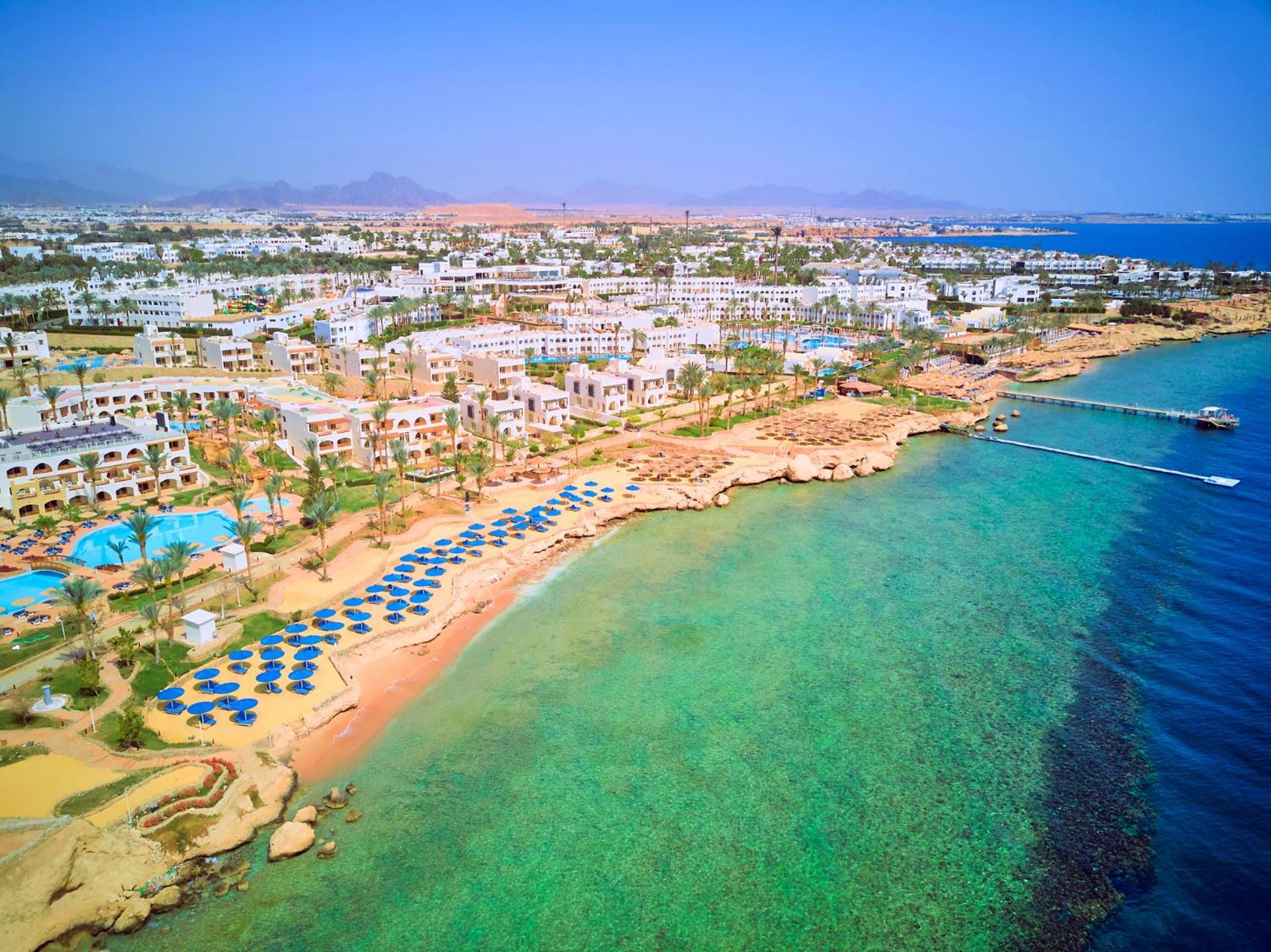 Pickalbatros Royal Grand Sharm - Adults Friendly 16 Years Plus Exteriér fotografie The photo showcases a picturesque coastal scene, likely from a resort area. It features a sandy beach lined with blue umbrellas and lounge chairs, indicating a recreational space for relaxation. The water is clear and exhibits shades of green and blu