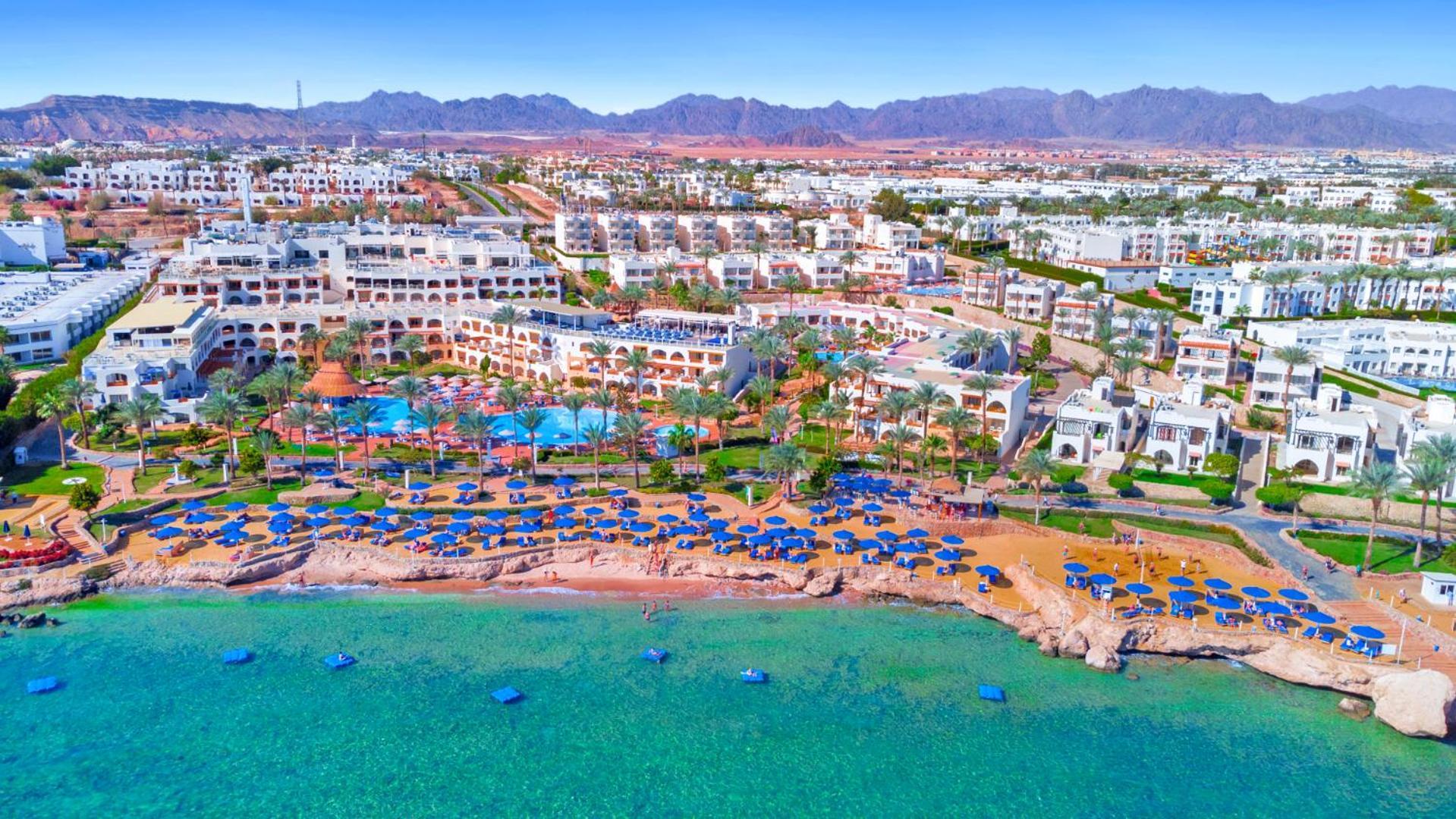 Pickalbatros Royal Grand Sharm - Adults Friendly 16 Years Plus Exteriér fotografie The photo showcases a beautiful coastal resort area. In the foreground, there is a sandy beach lined with blue umbrellas and sun loungers. The vibrant turquoise water gently laps at the shore. Behind the beach, you can see a well-maintained resort wi