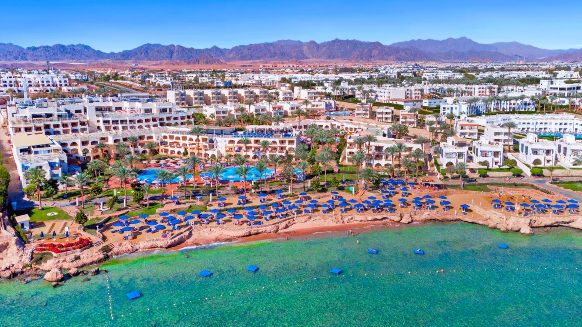 Pickalbatros Royal Grand Sharm - Adults Friendly 16 Years Plus Exteriér fotografie The photo shows an aerial view of a coastal resort area. In the foreground, there is a beach with greenish-blue water, where several blue beach umbrellas and lounges are set up for guests. The shoreline appears sandy, and there are some rocky areas. 