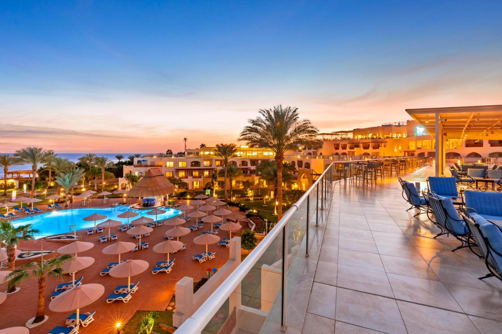 Pickalbatros Royal Grand Sharm - Adults Friendly 16 Years Plus Exteriér fotografie The photo depicts a scenic view of a resort during sunset. In the foreground, there is a balcony with tiled flooring, overlooking the resort. Below, a swimming pool is visible, surrounded by lounge chairs and large umbrellas, indicating a relaxing at