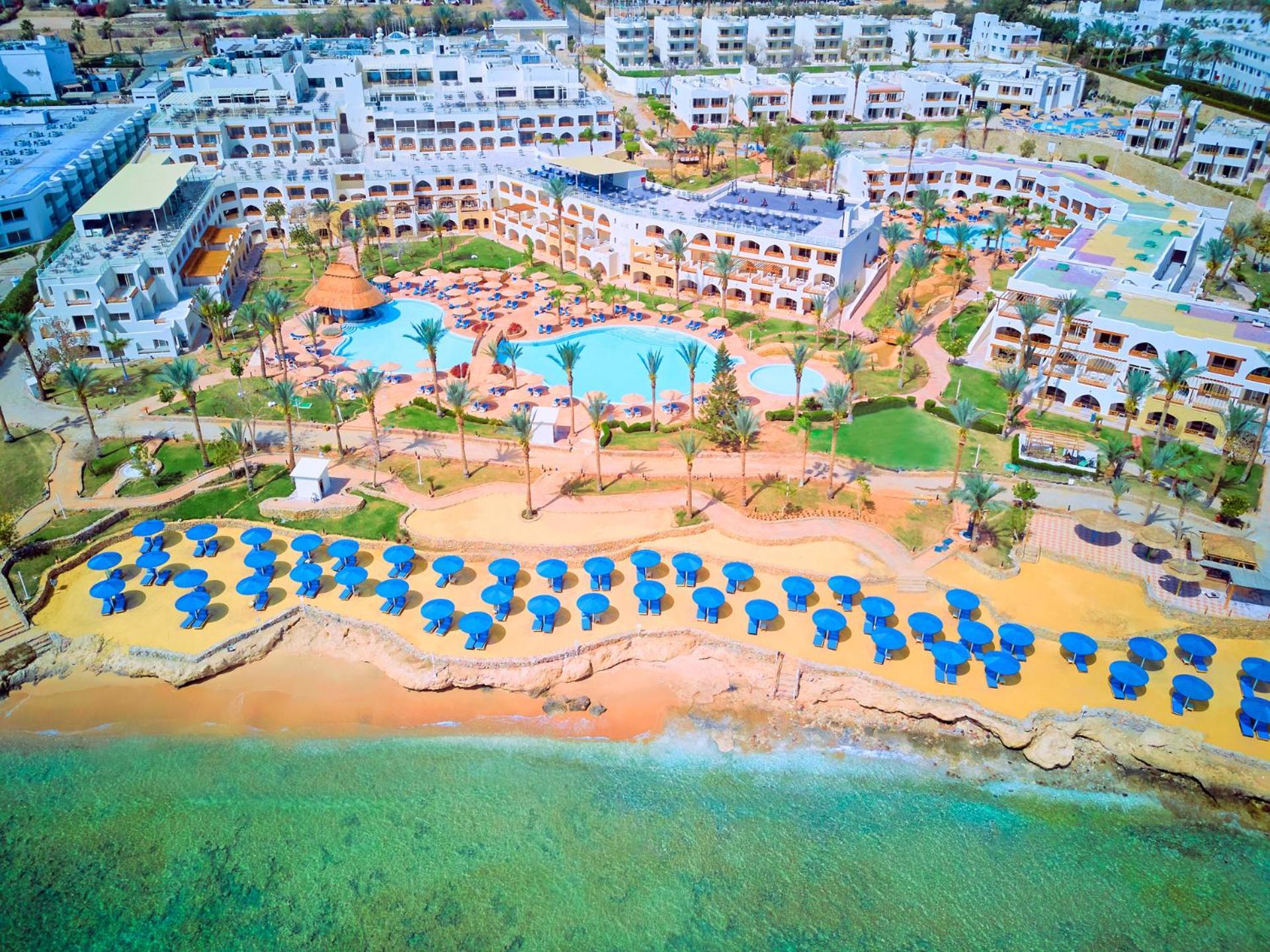 Pickalbatros Royal Grand Sharm - Adults Friendly 16 Years Plus Exteriér fotografie The photo shows a large resort complex situated near a beach. There is a turquoise swimming pool at the center, surrounded by green lawns and palm trees. Sun loungers with blue umbrellas are lined up on the sandy beach, and the beach area is adjacent