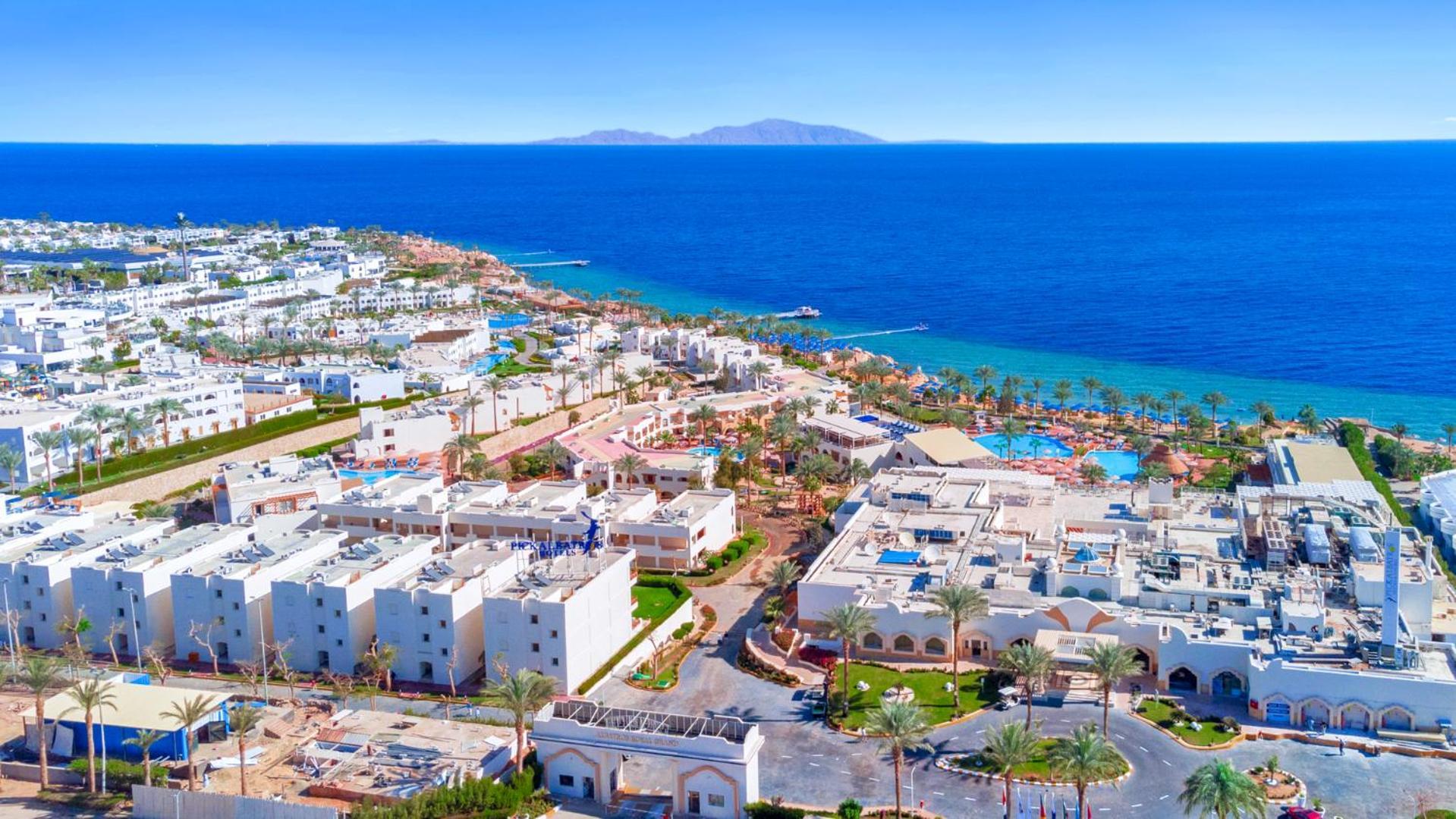 Pickalbatros Royal Grand Sharm - Adults Friendly 16 Years Plus Exteriér fotografie The photo shows a beautiful coastal landscape, featuring a resort area by the sea. There are white buildings, likely hotels or villas, arranged in a sprawling layout near the beach. The water is a vibrant blue, contrasting with the sandy shore and gr