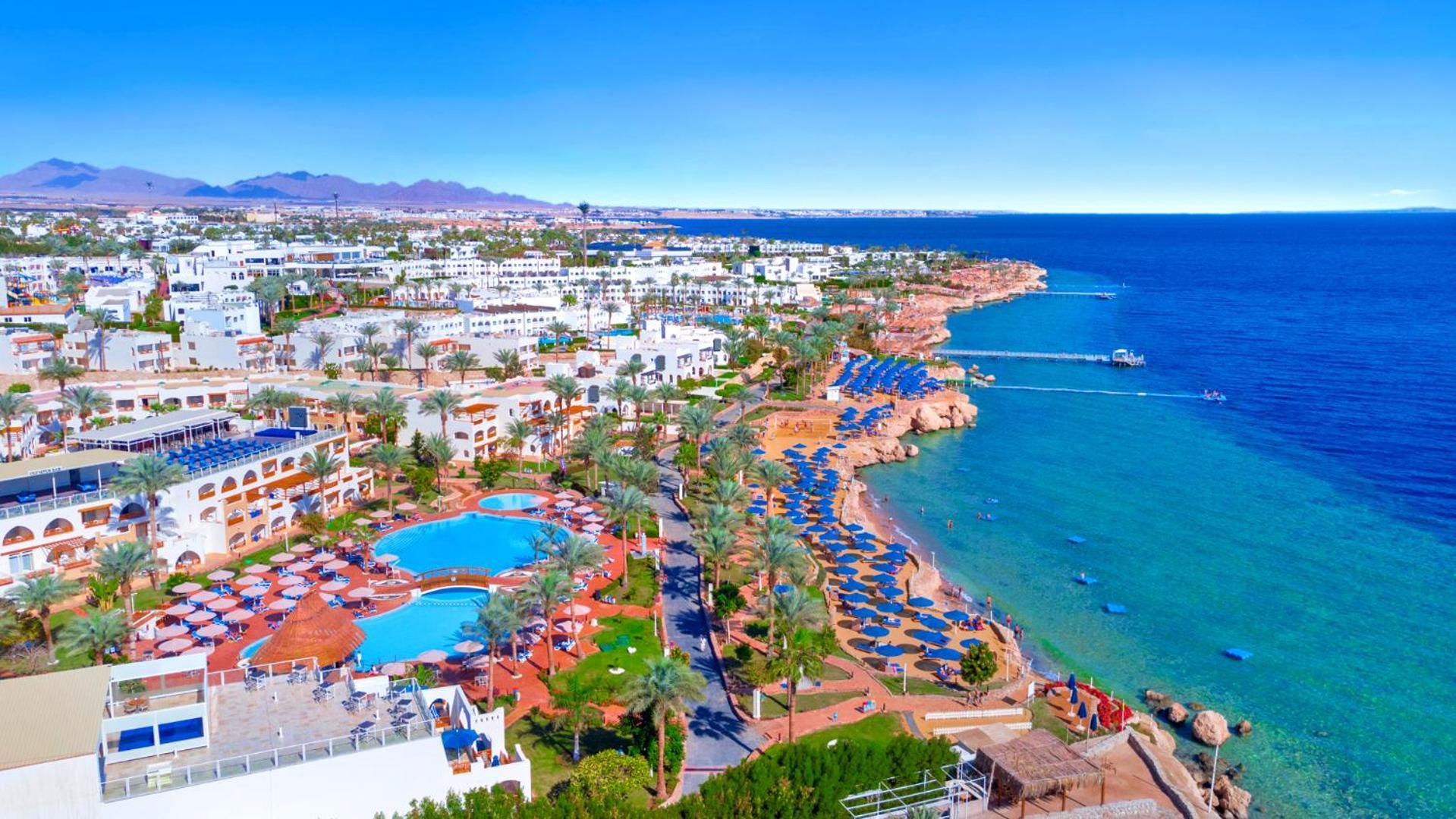 Pickalbatros Royal Grand Sharm - Adults Friendly 16 Years Plus Exteriér fotografie The photo features a picturesque coastal scene, likely from a resort area. In the foreground, there is a large swimming pool surrounded by lounge chairs. Further back, the landscape includes several buildings, possibly hotels or apartments, that over