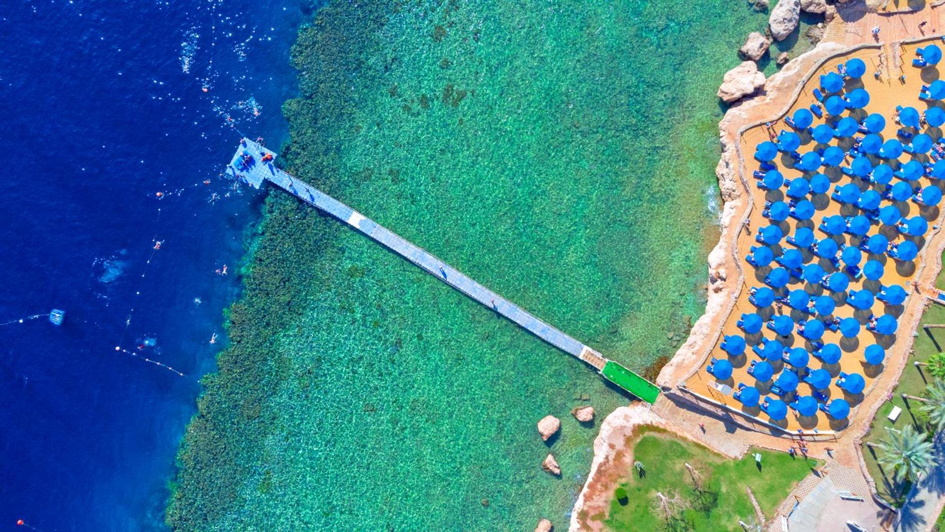 Pickalbatros Royal Grand Sharm - Adults Friendly 16 Years Plus Exteriér fotografie The photo shows an aerial view of a coastal area featuring a long wooden pier extending into clear turquoise water. On one side of the pier, there appears to be a beach with umbrellas or sunbeds arranged neatly. The water is vibrant and visually invi