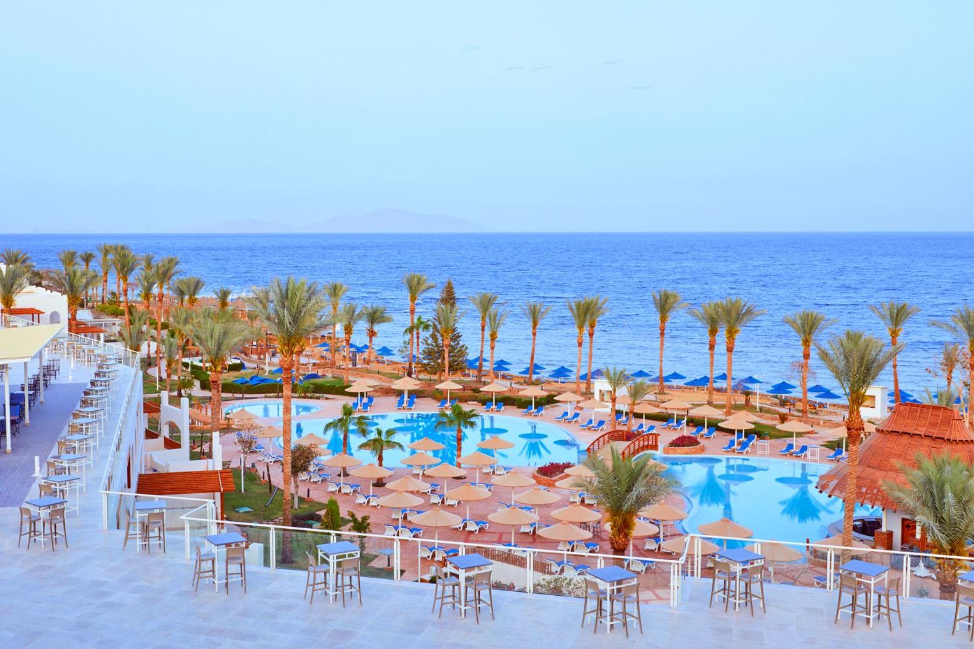 Pickalbatros Royal Grand Sharm - Adults Friendly 16 Years Plus Exteriér fotografie The photo depicts a scenic resort area by the sea. In the foreground, there is a large swimming pool surrounded by lounge chairs and umbrellas. Palm trees line the sides of the pool, adding to the tropical atmosphere. In the background, a beautiful b