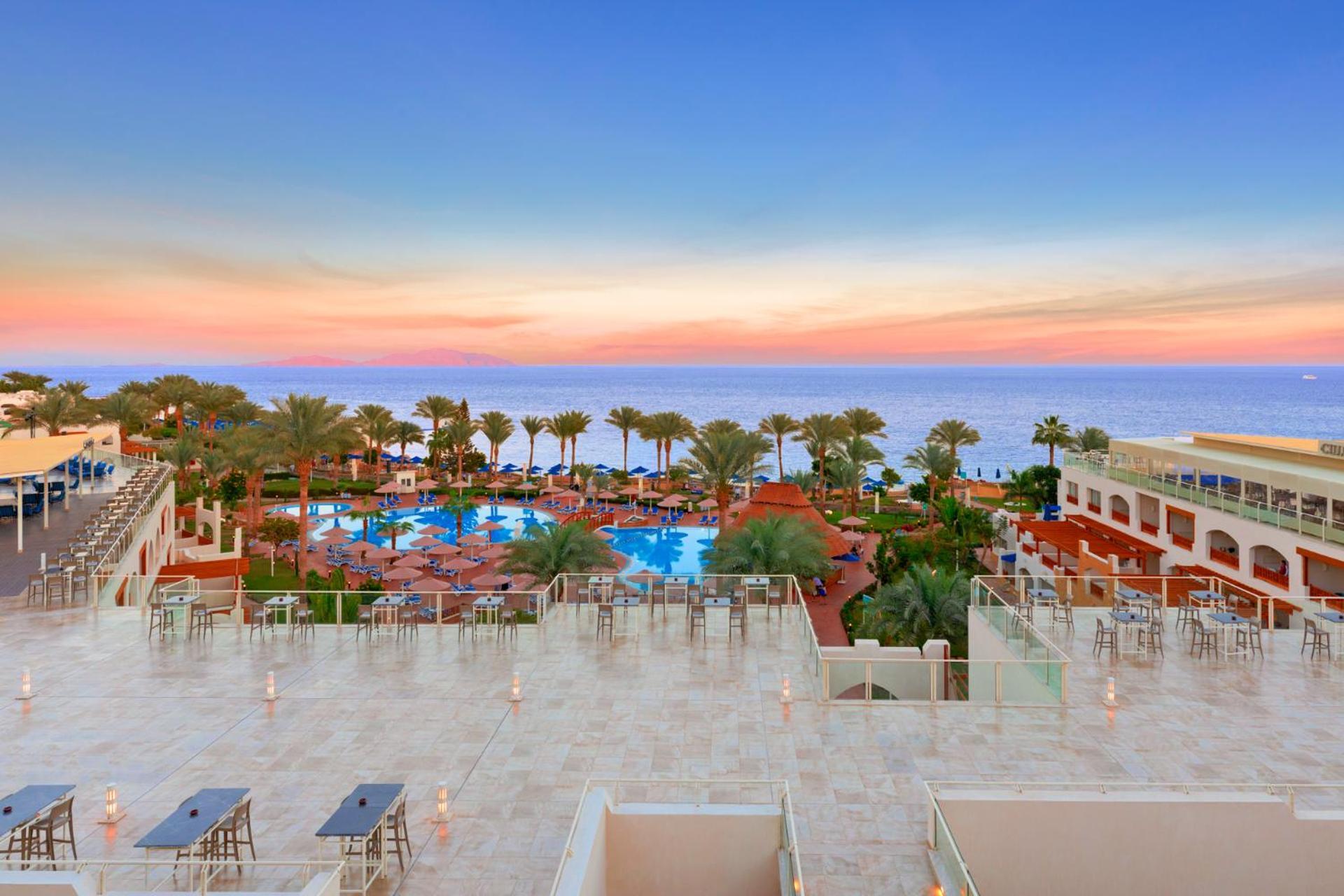 Pickalbatros Royal Grand Sharm - Adults Friendly 16 Years Plus Exteriér fotografie The photo depicts a beautiful resort scene at sunset. In the foreground, there is a spacious terrace area with modern furniture. Below, a large swimming pool is surrounded by lush palm trees and lounge chairs, indicating a relaxing atmosphere. The vi