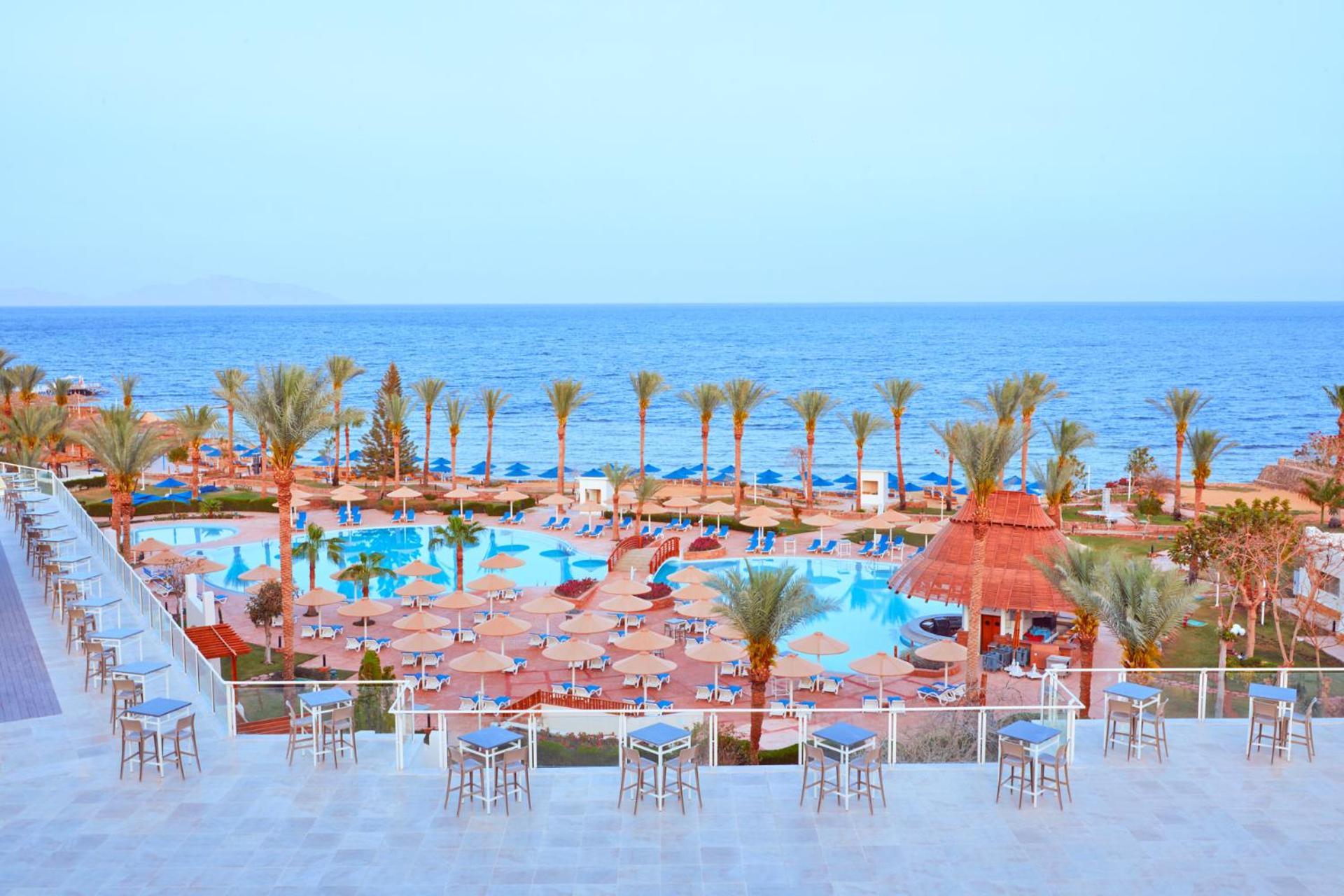 Pickalbatros Royal Grand Sharm - Adults Friendly 16 Years Plus Exteriér fotografie The photo shows a scenic view of a resort or beach area. In the foreground, there are several seating areas with bar stools on a terrace. Below, a large swimming pool is visible, surrounded by lounging chairs and umbrellas. Palm trees line the area, 