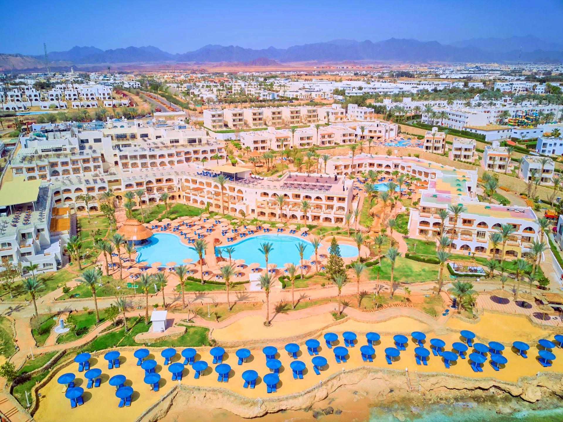 Pickalbatros Royal Grand Sharm - Adults Friendly 16 Years Plus Exteriér fotografie The photo shows a large resort complex set in a scenic location, likely near a beach. The layout features multiple buildings with a Mediterranean style of architecture, surrounded by lush greenery and palm trees. Central to the resort is a swimming p