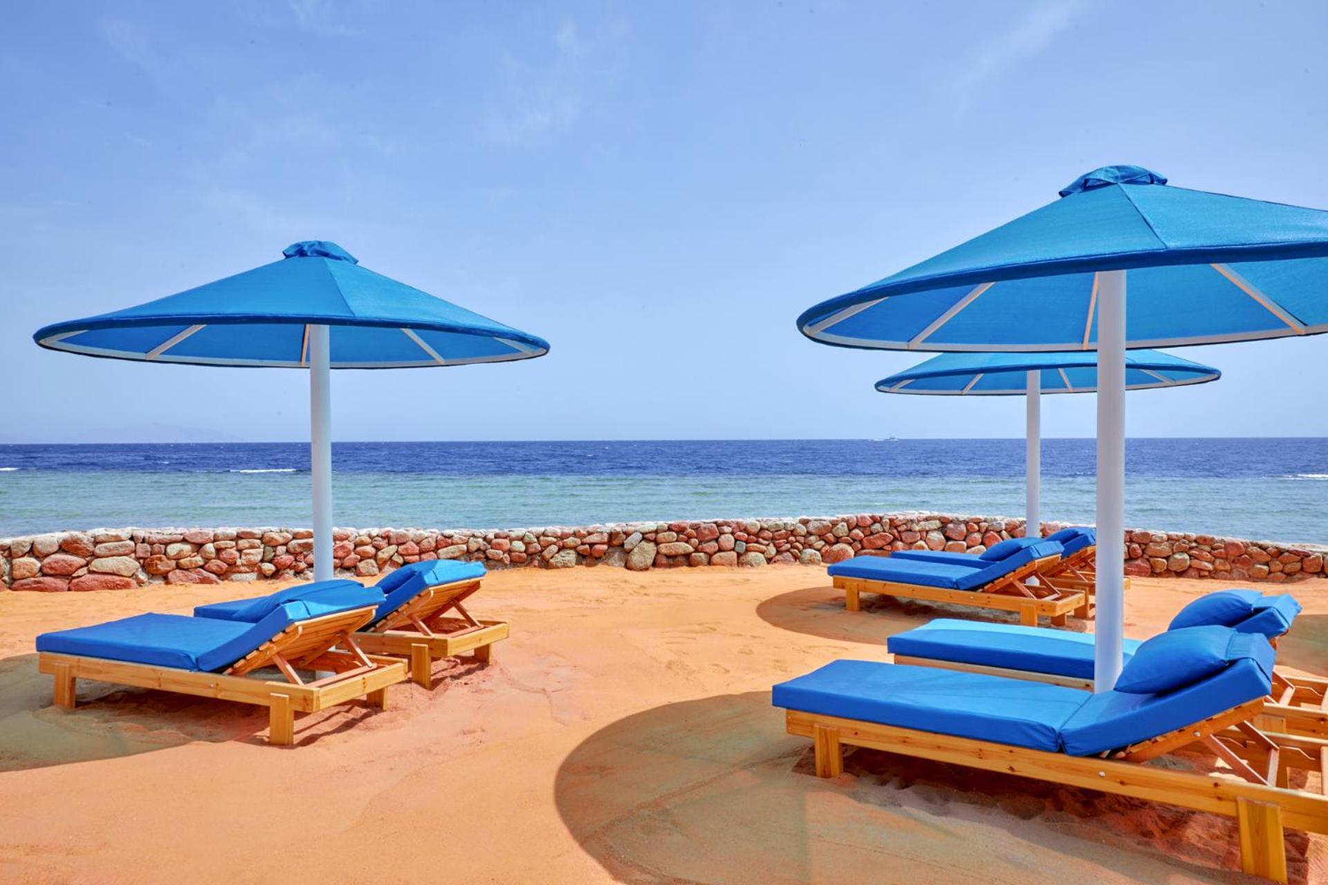 Pickalbatros Royal Grand Sharm - Adults Friendly 16 Years Plus Exteriér fotografie The photo shows a beach scene featuring several blue umbrellas and lounge chairs on a sandy area. The umbrellas are large and have a bright blue color, providing shade for the lounge chairs underneath. In the background, there's a view of the ocean w