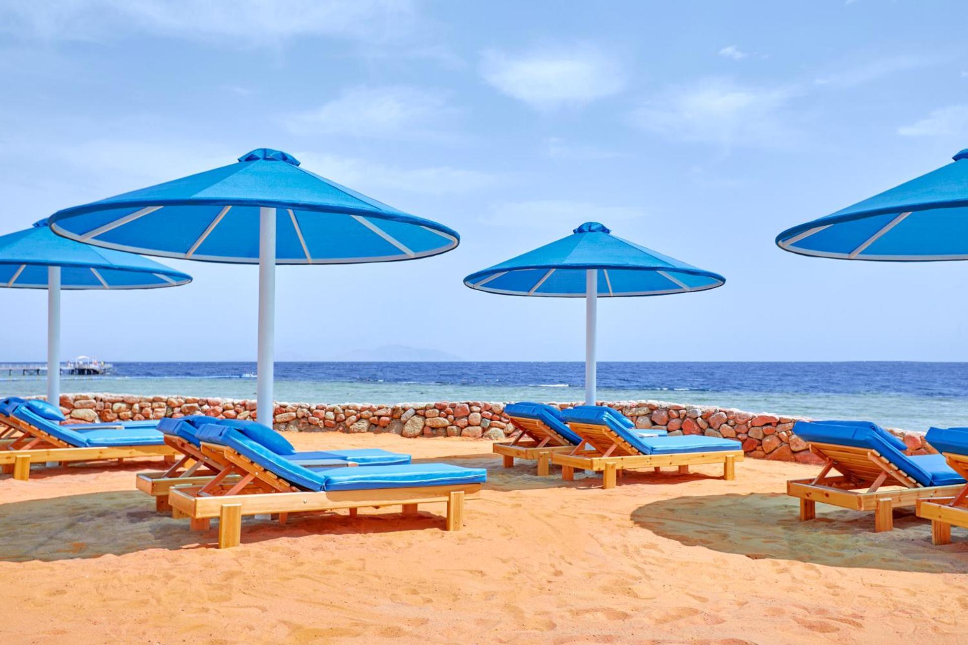 Pickalbatros Royal Grand Sharm - Adults Friendly 16 Years Plus Exteriér fotografie The photo shows a beach scene featuring several blue umbrellas providing shade over wooden lounge chairs. The sandy beach extends toward a calm sea, creating a relaxing atmosphere. The sky is mostly clear, with a few clouds, suggesting pleasant weath