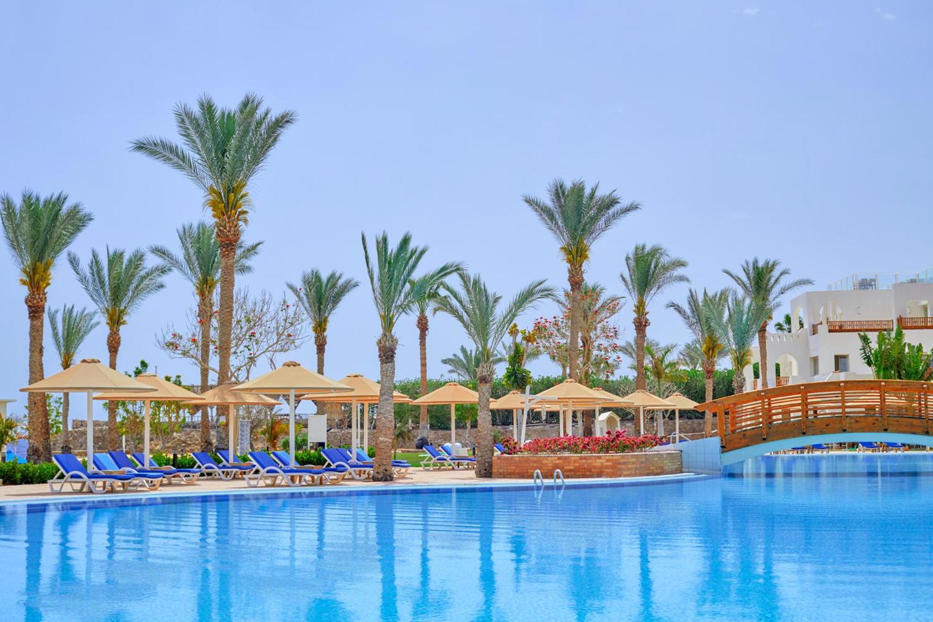 Pickalbatros Royal Grand Sharm - Adults Friendly 16 Years Plus Exteriér fotografie The photo shows a serene poolside scene at a resort. There is a large, clear blue pool reflecting the sky, surrounded by lounge chairs with blue cushions. Palm trees line the area, adding a tropical feel. Several covered loungers are placed under umb