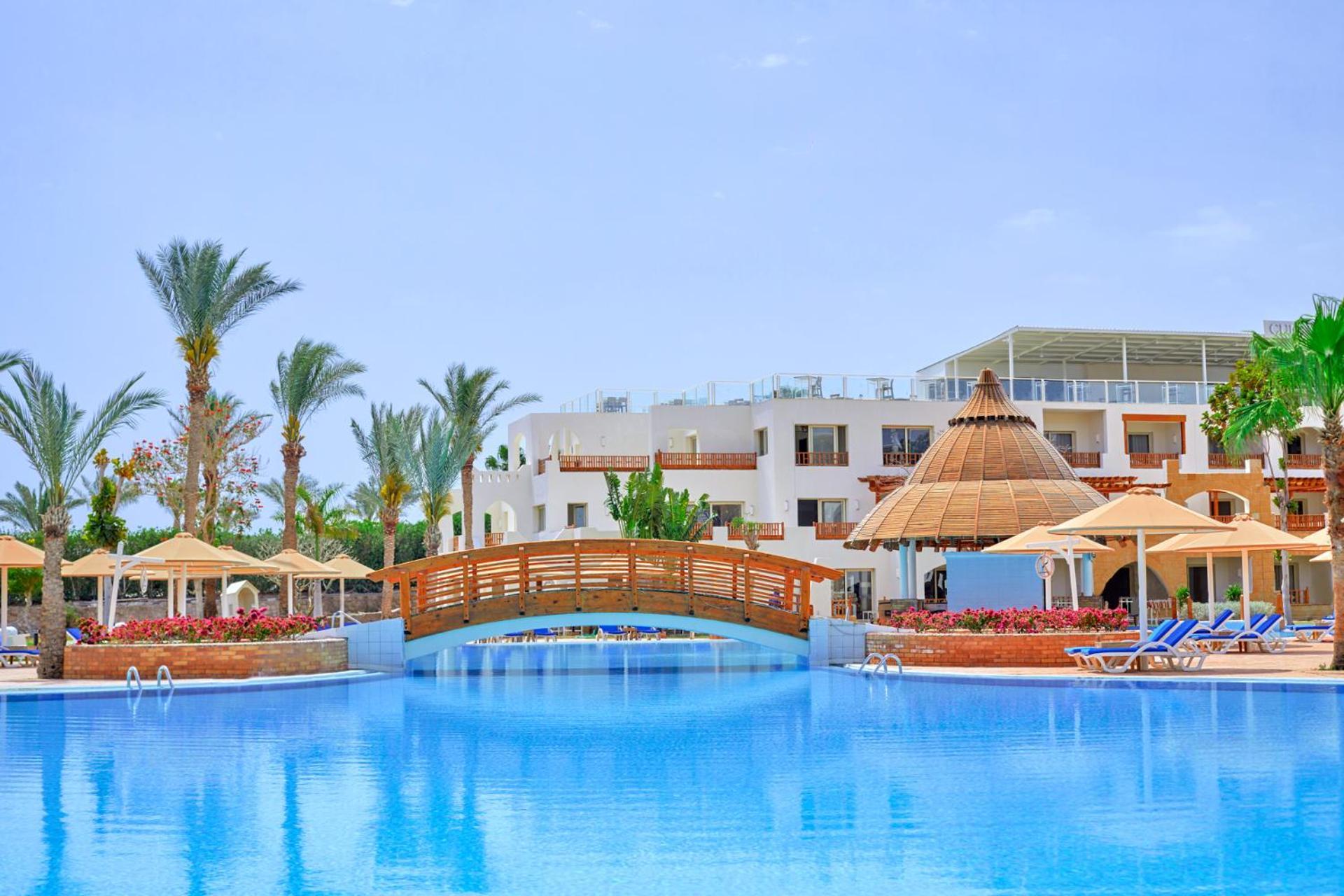 Pickalbatros Royal Grand Sharm - Adults Friendly 16 Years Plus Exteriér fotografie The photo depicts a serene resort setting featuring a large, calm swimming pool, which reflects the clear blue sky. In the foreground, a wooden bridge arches over the pool. Surrounding the pool are several palm trees and tropical foliage, contributin