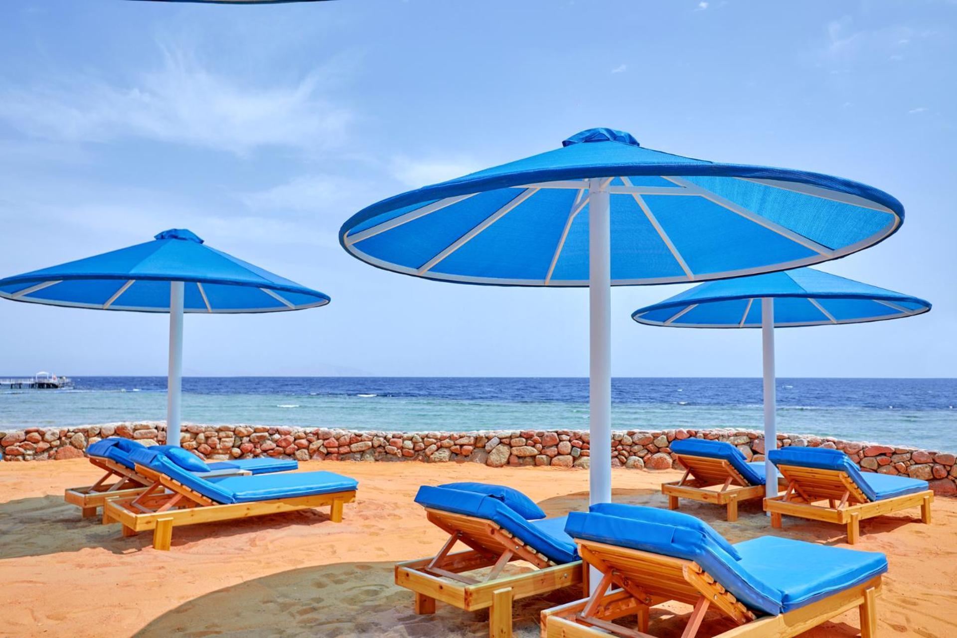 Pickalbatros Royal Grand Sharm - Adults Friendly 16 Years Plus Exteriér fotografie The photo shows a serene beach scene with several blue sun loungers arranged beneath large blue umbrellas. The loungers are positioned on sandy ground, with the ocean visible in the background. The sky is clear, indicating a bright and sunny day, and