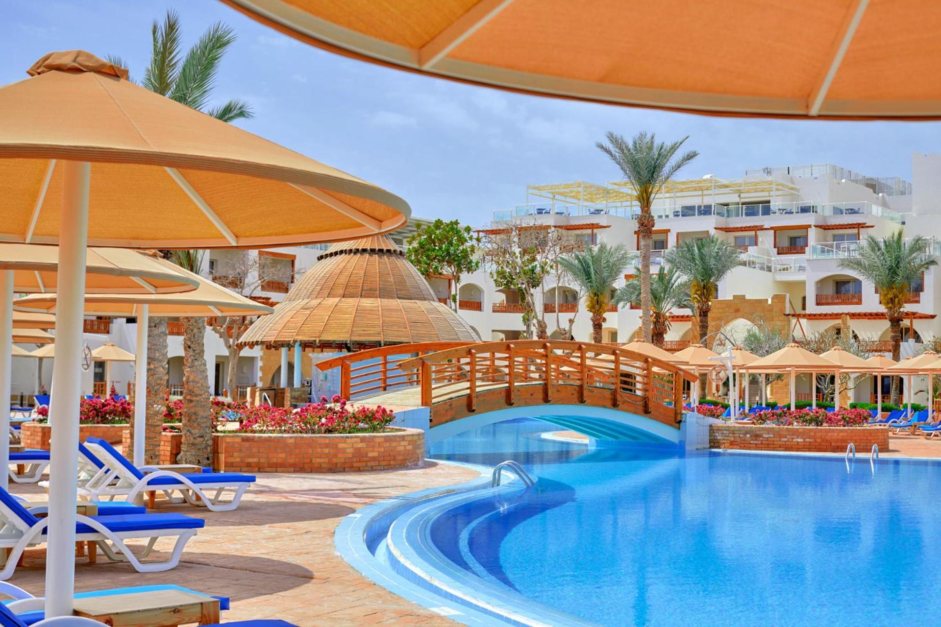 Pickalbatros Royal Grand Sharm - Adults Friendly 16 Years Plus Exteriér fotografie The photo shows a picturesque resort scene featuring a bright blue swimming pool surrounded by sun loungers and parasols. In the background, there is a wooden bridge arching over a section of the pool, leading towards the other side. The area is ador