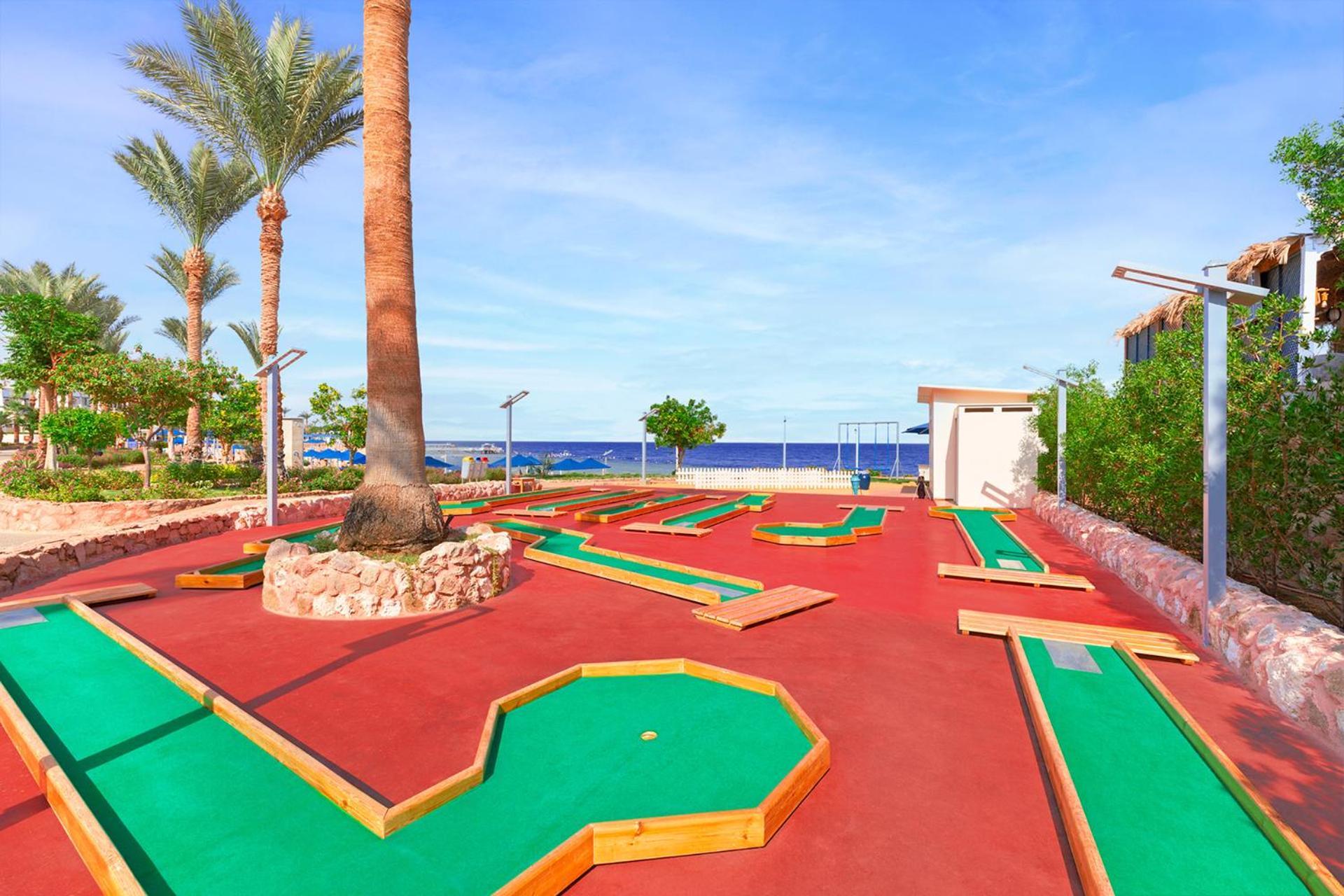 Pickalbatros Royal Grand Sharm - Adults Friendly 16 Years Plus Exteriér fotografie The photo shows a mini-golf course situated outdoors, with a view of the sea in the background. The course features vibrant green and red surfaces, laid out in winding patterns, and surrounded by well-maintained landscaping, including palm trees. The