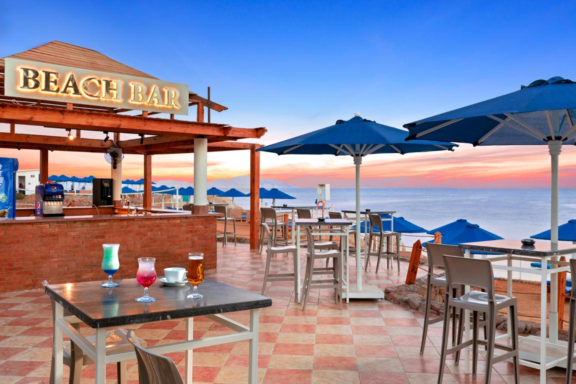 Pickalbatros Royal Grand Sharm - Adults Friendly 16 Years Plus Exteriér fotografie The photo shows a beach bar setting, featuring a wooden structure with the words "BEACH BAR" prominently displayed. There are several tables and chairs arranged on a tiled floor, with some drinks placed on the tables. Blue umbrellas provide shade for