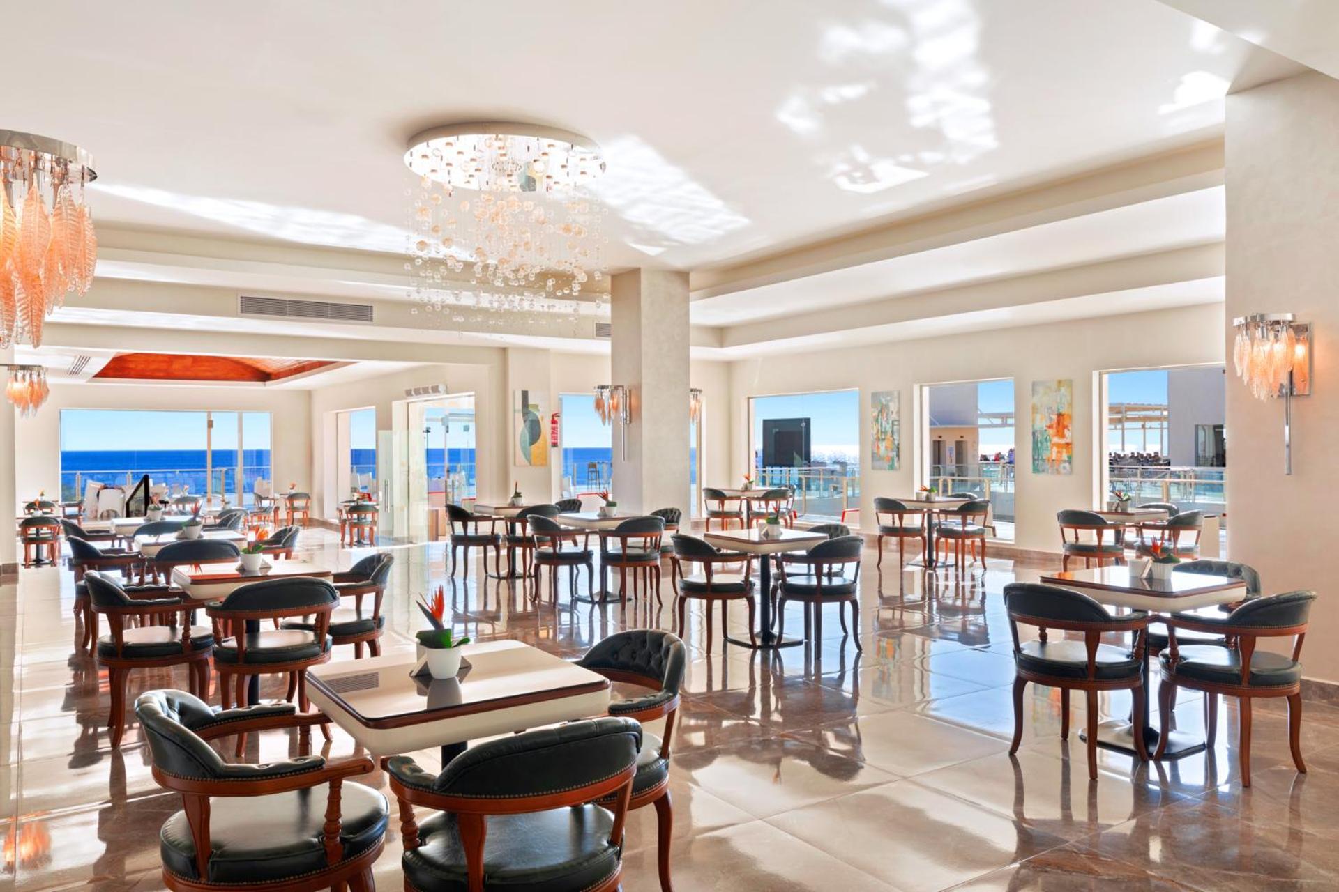 Pickalbatros Royal Grand Sharm - Adults Friendly 16 Years Plus Exteriér fotografie The photo shows a bright and elegant restaurant interior. There are several tables arranged throughout the space, each with chairs around them. The decor features modern lighting, including a chandelier, and large windows that allow natural light to 