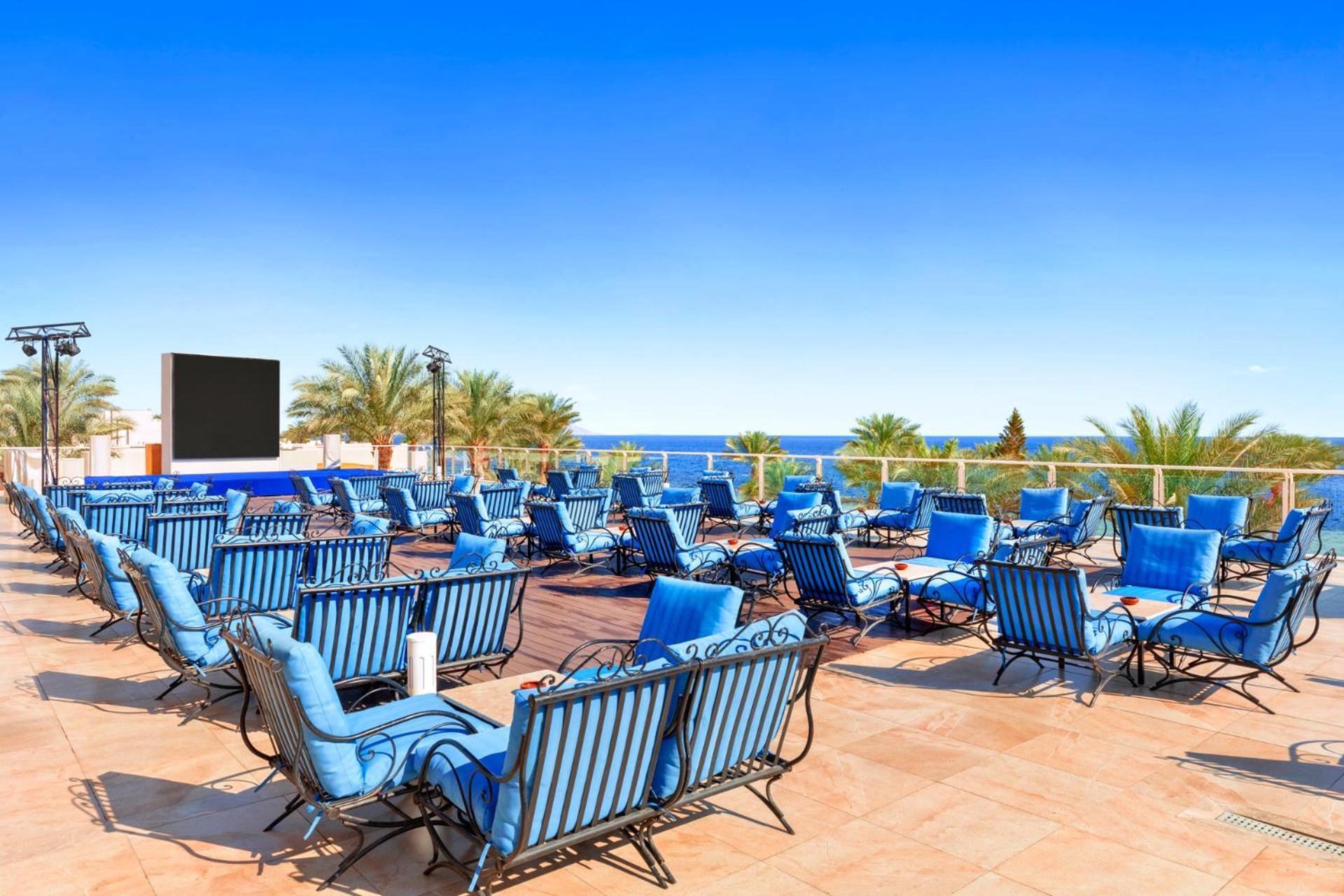 Pickalbatros Royal Grand Sharm - Adults Friendly 16 Years Plus Exteriér fotografie The photo features a sunny outdoor space with numerous blue lounge chairs arranged in rows on a tiled patio. In the background, there is a clear blue sky and a view of the ocean, along with some palm trees adding to the tropical atmosphere. Additiona
