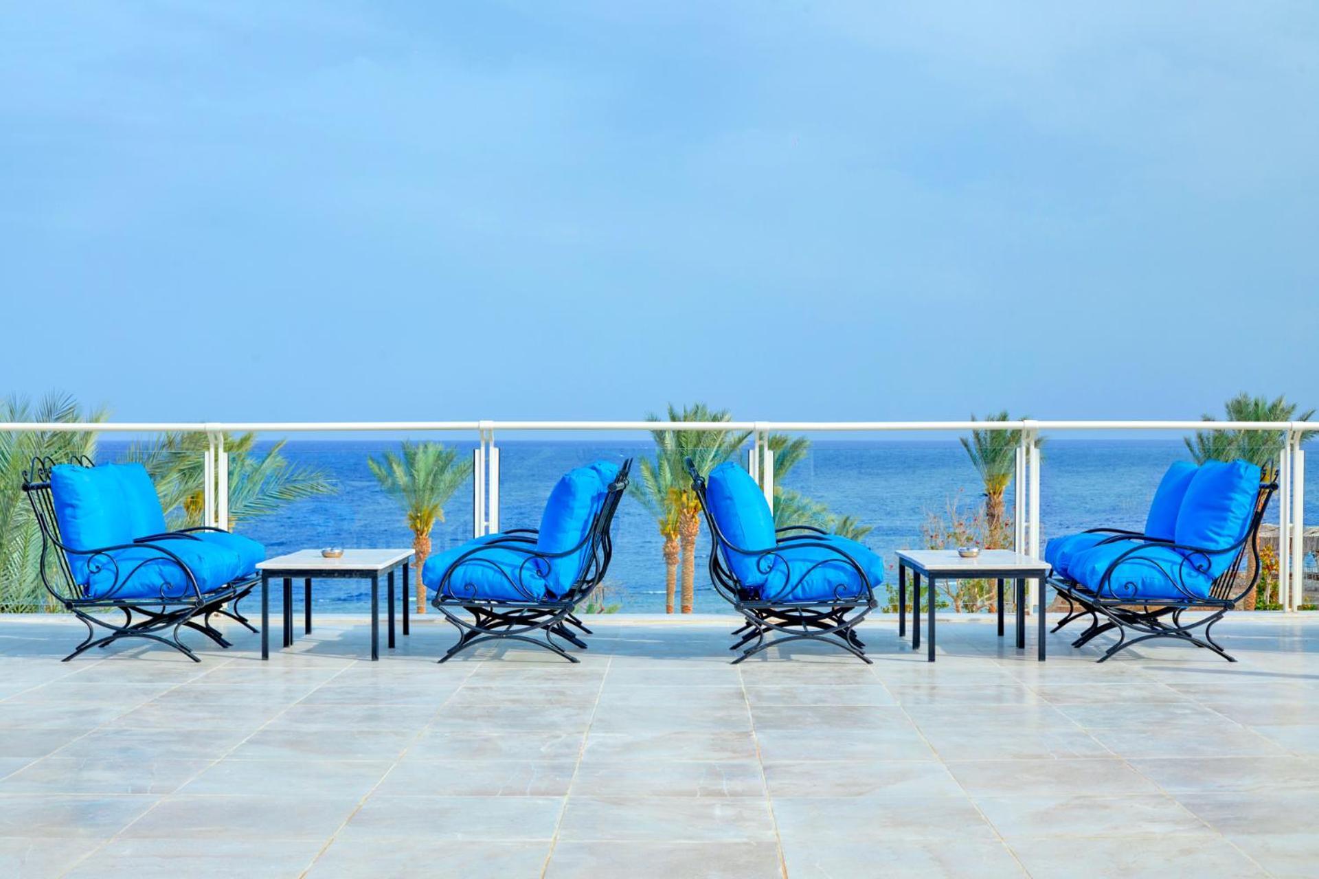 Pickalbatros Royal Grand Sharm - Adults Friendly 16 Years Plus Exteriér fotografie The photo depicts a serene outdoor patio area with four blue lounge chairs arranged around small tables. The setting features a clear view of the ocean with palm trees visible in the background, complementing the tranquil atmosphere. The overall colo