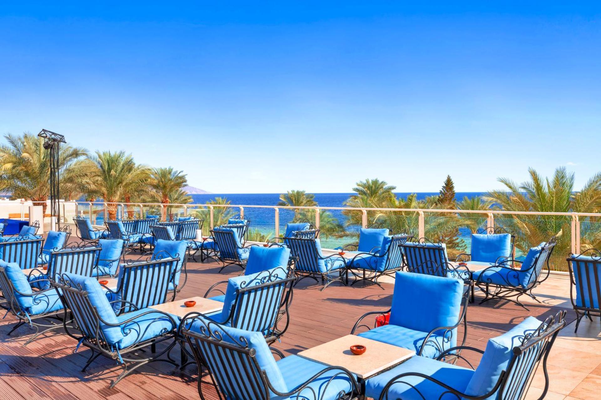 Pickalbatros Royal Grand Sharm - Adults Friendly 16 Years Plus Exteriér fotografie The photo shows an outdoor deck or terrace area, adorned with numerous blue lounge chairs arranged neatly. In the background, there are palm trees and a clear blue sea that stretches toward the horizon under a bright sky. The setting appears to be a 