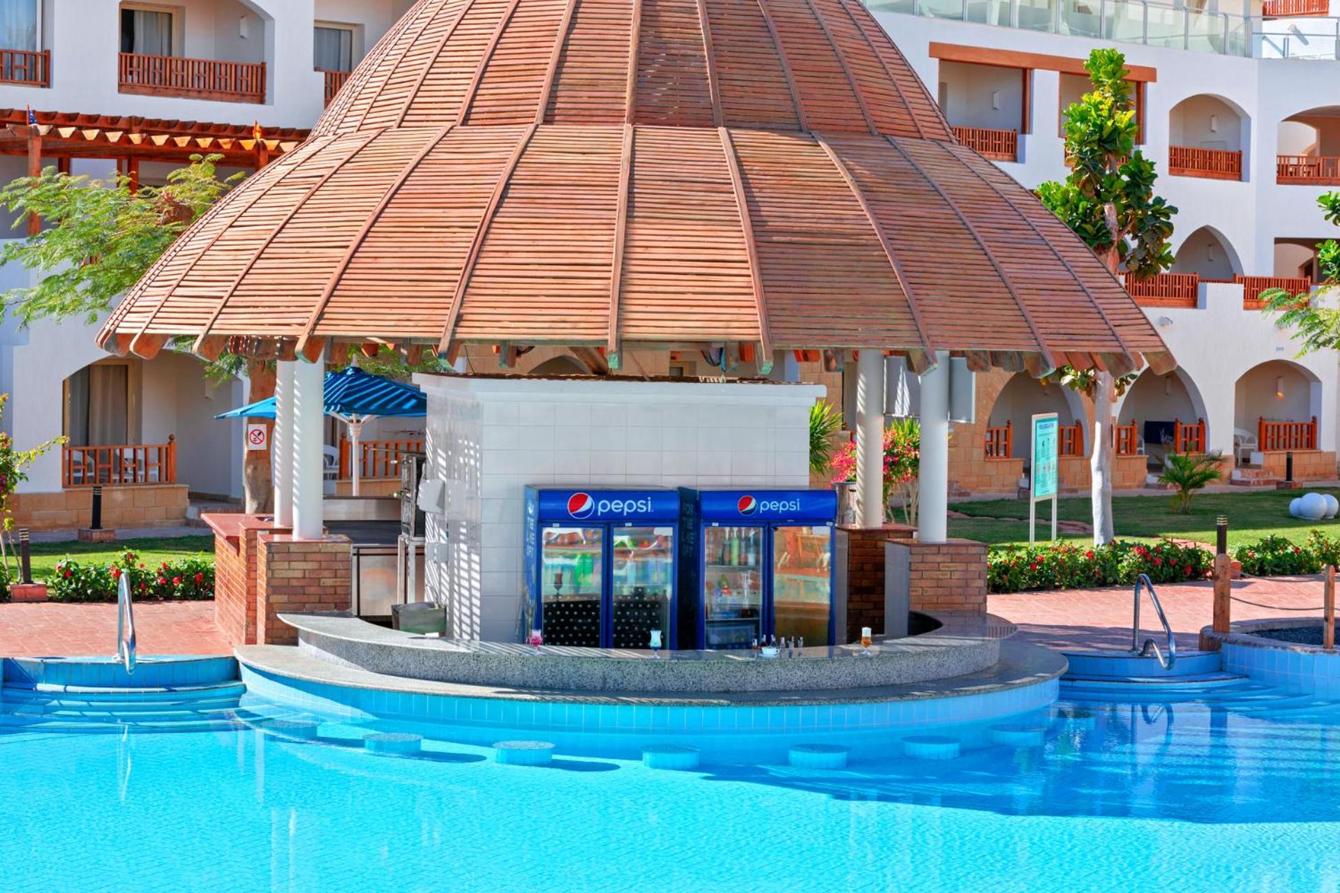 Pickalbatros Royal Grand Sharm - Adults Friendly 16 Years Plus Exteriér fotografie The photo shows a poolside bar with a thatched roof structure, located near a swimming pool. The bar features a counter where drinks are served, and there are vending machines displaying the Pepsi brand. Surrounding the bar, there are neatly landscap